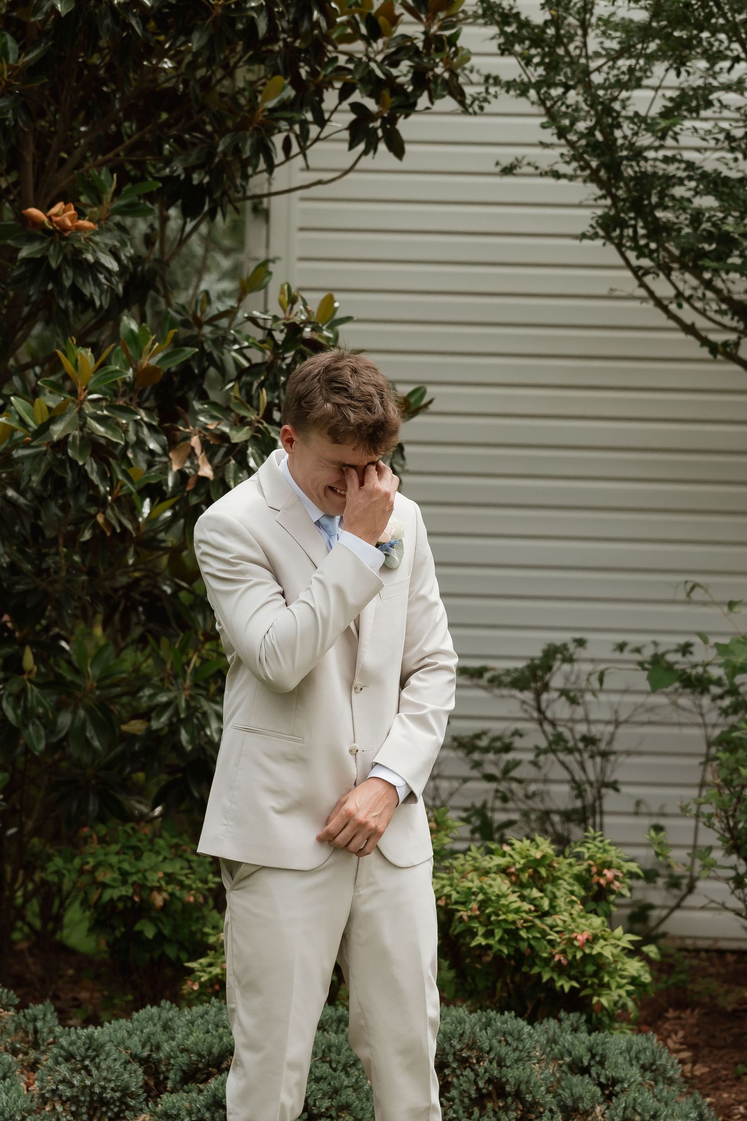 Emotional groom in beige suit wiping tears outdoors among lush greenery before wedding ceremony.