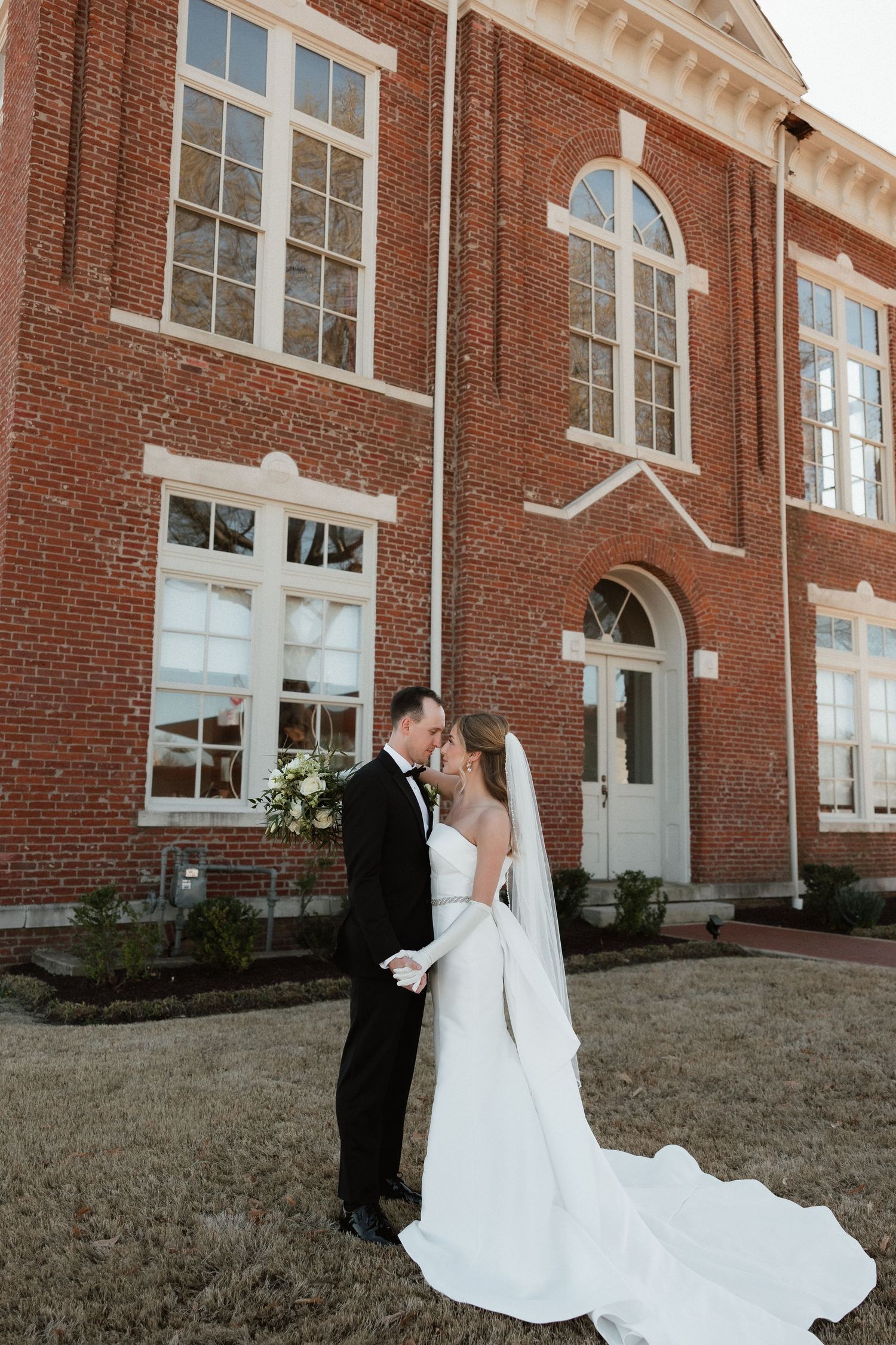 Bride and groom share a romantic nose-to-nose moment on the lawn of a grand brick building.