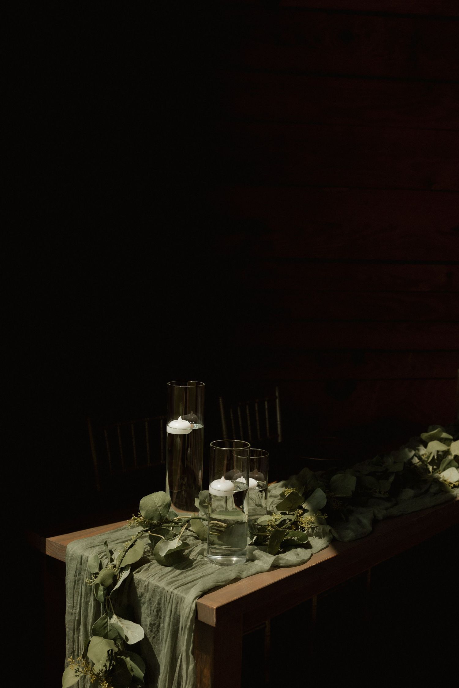 Rustic wedding reception table with candles in glass holders and eucalyptus garland in dark setting.