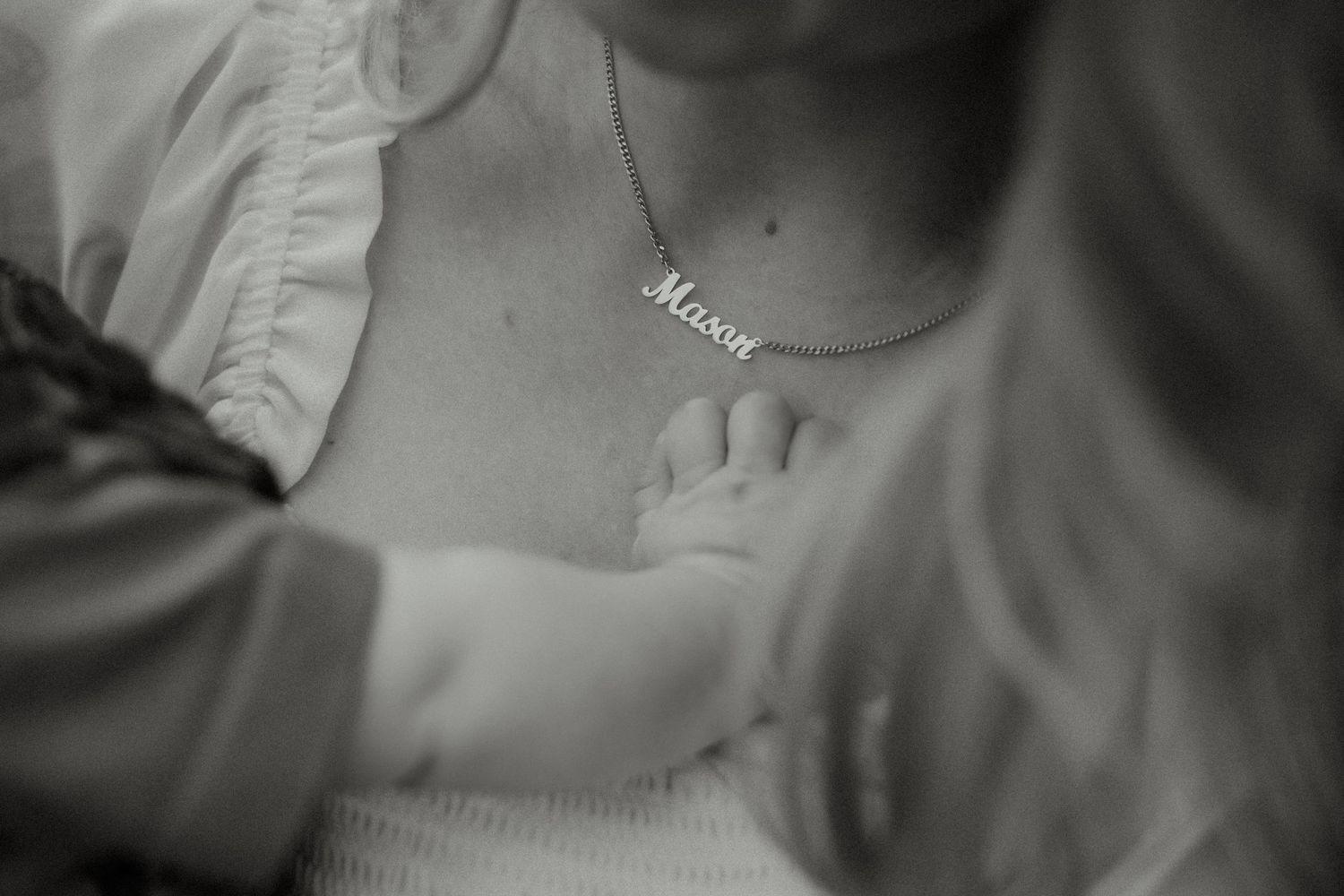 Black and white close-up of baby touching mothers chest wearing a delicate name necklace.