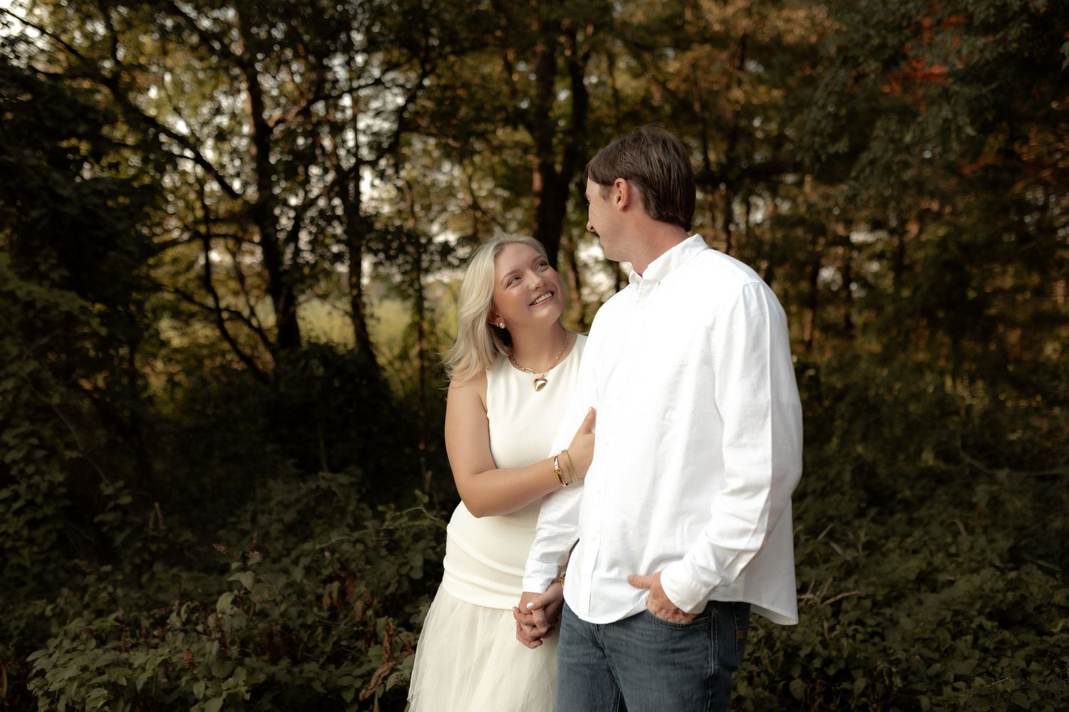 Couple holding hands and smiling at each other in a lush green forest setting.