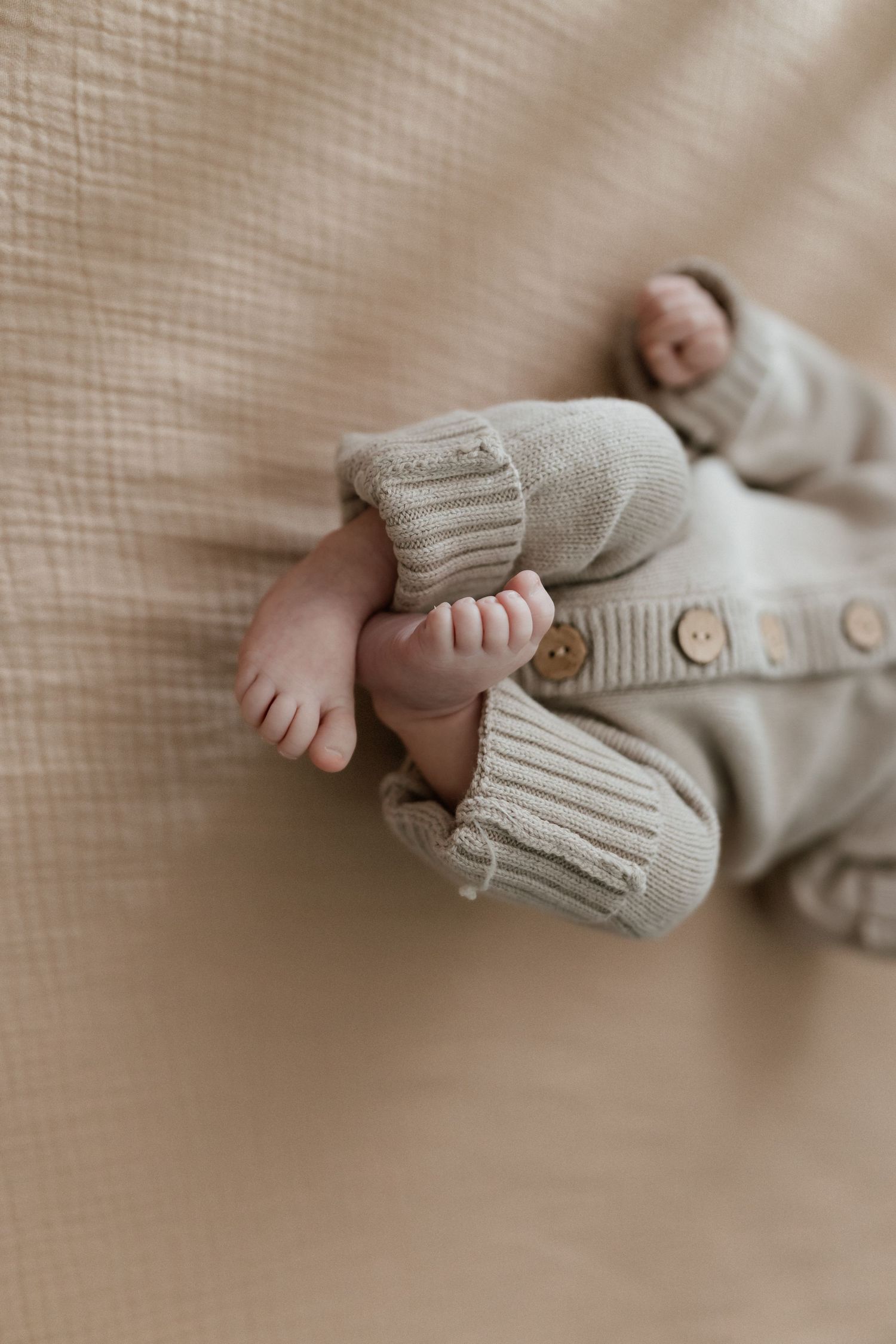 Close-up of a newborn baby\u2019s tiny feet in a sage green knit romper lying on a soft beige blanket.
