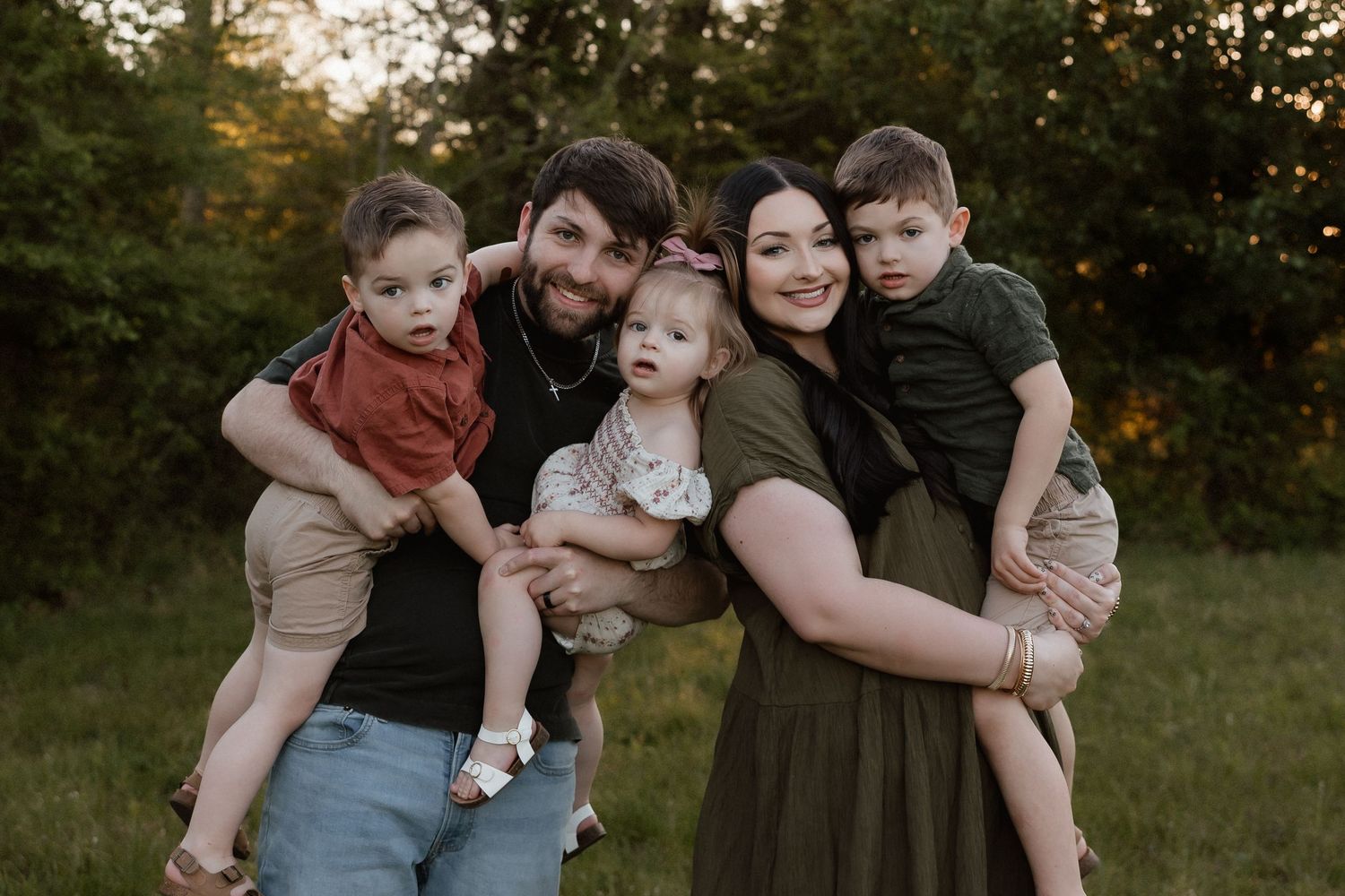 Happy family of five posing together outdoors at sunset, parents holding three young children in a lush green setting.