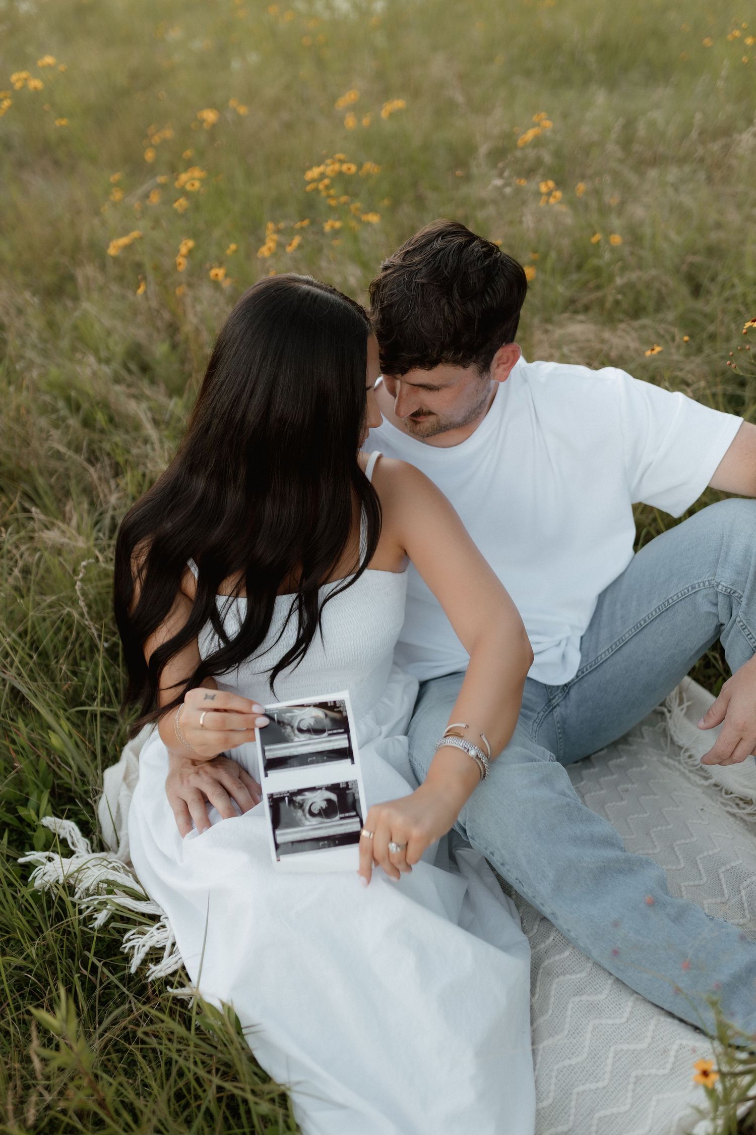 Expecting couple sitting in a meadow holding ultrasound photos, woman in white dress with long dark hair, golden hour light.