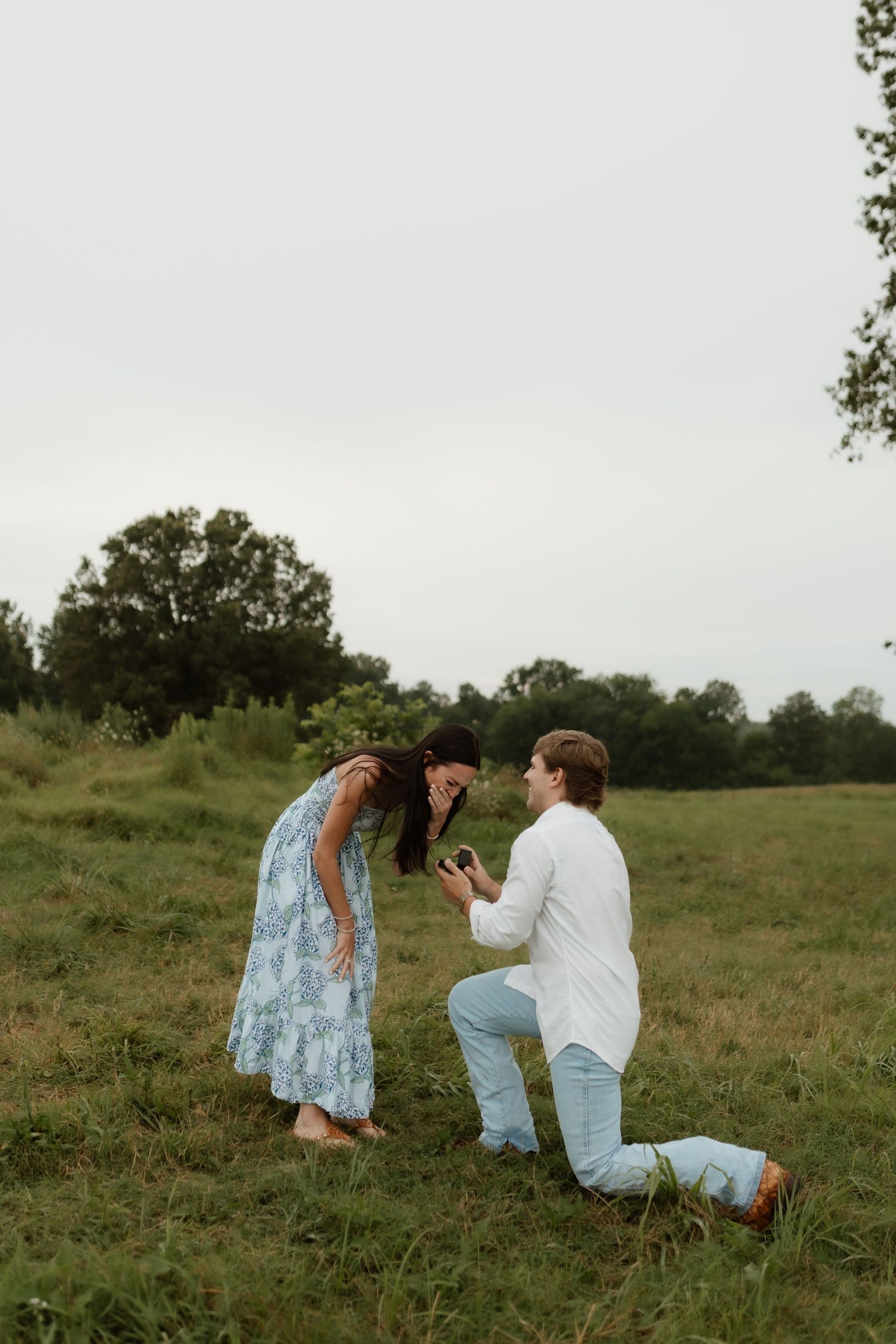 A man kneels on one knee proposing to a surprised woman in a blue floral dress in a green field.