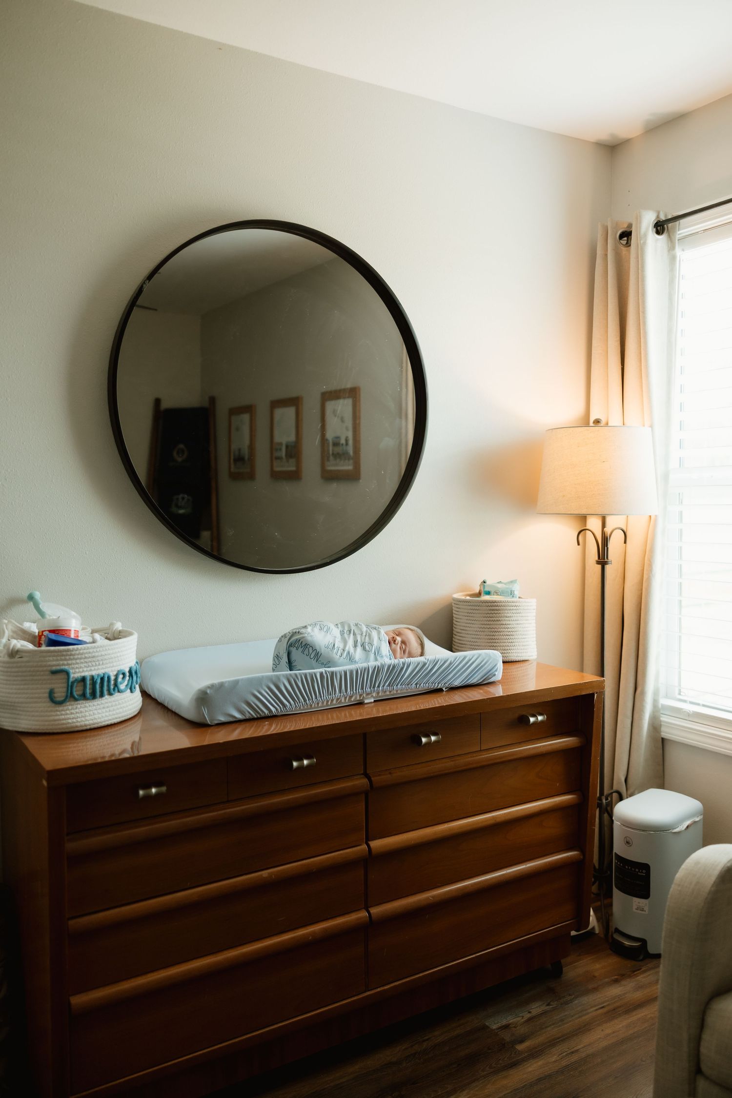 Wooden dresser with round mirror above changing pad holding newborn baby in softly lit nursery.