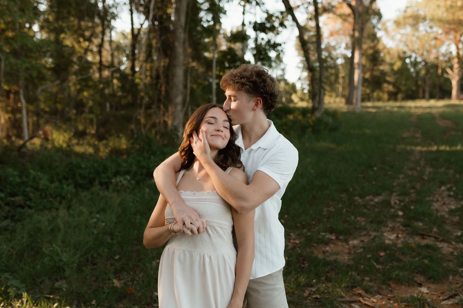 Young couple embracing in a sunlit forest, woman in white dress smiling as man holds her from behind.