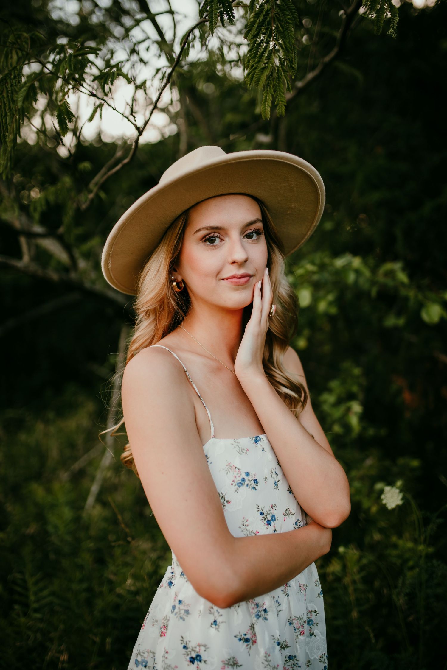 Young blonde woman in floral sundress and tan wide-brim hat poses gracefully among lush green foliage outdoors.