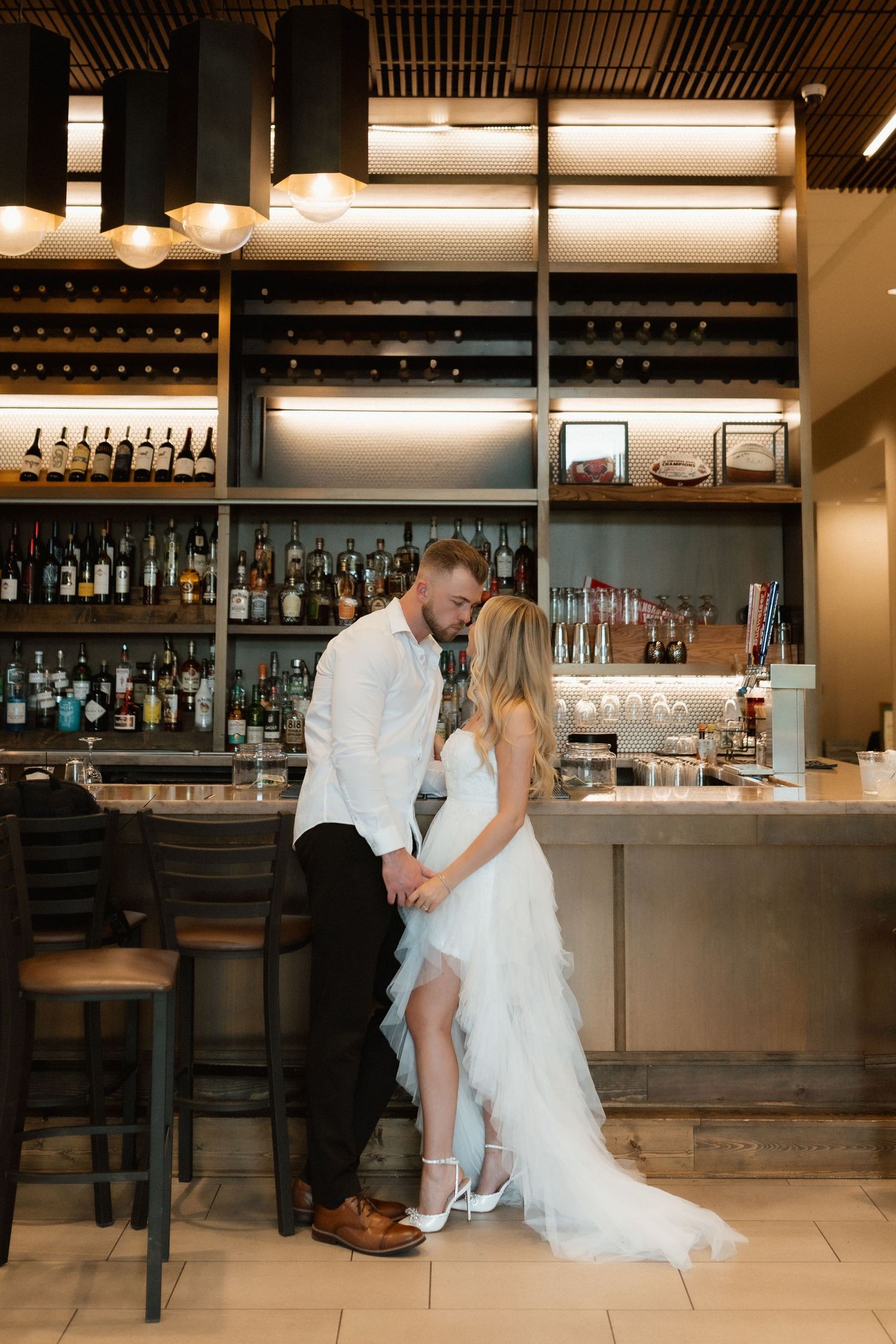 Bride and groom sharing romantic moment at elegant bar with wine bottle display in wedding venue.