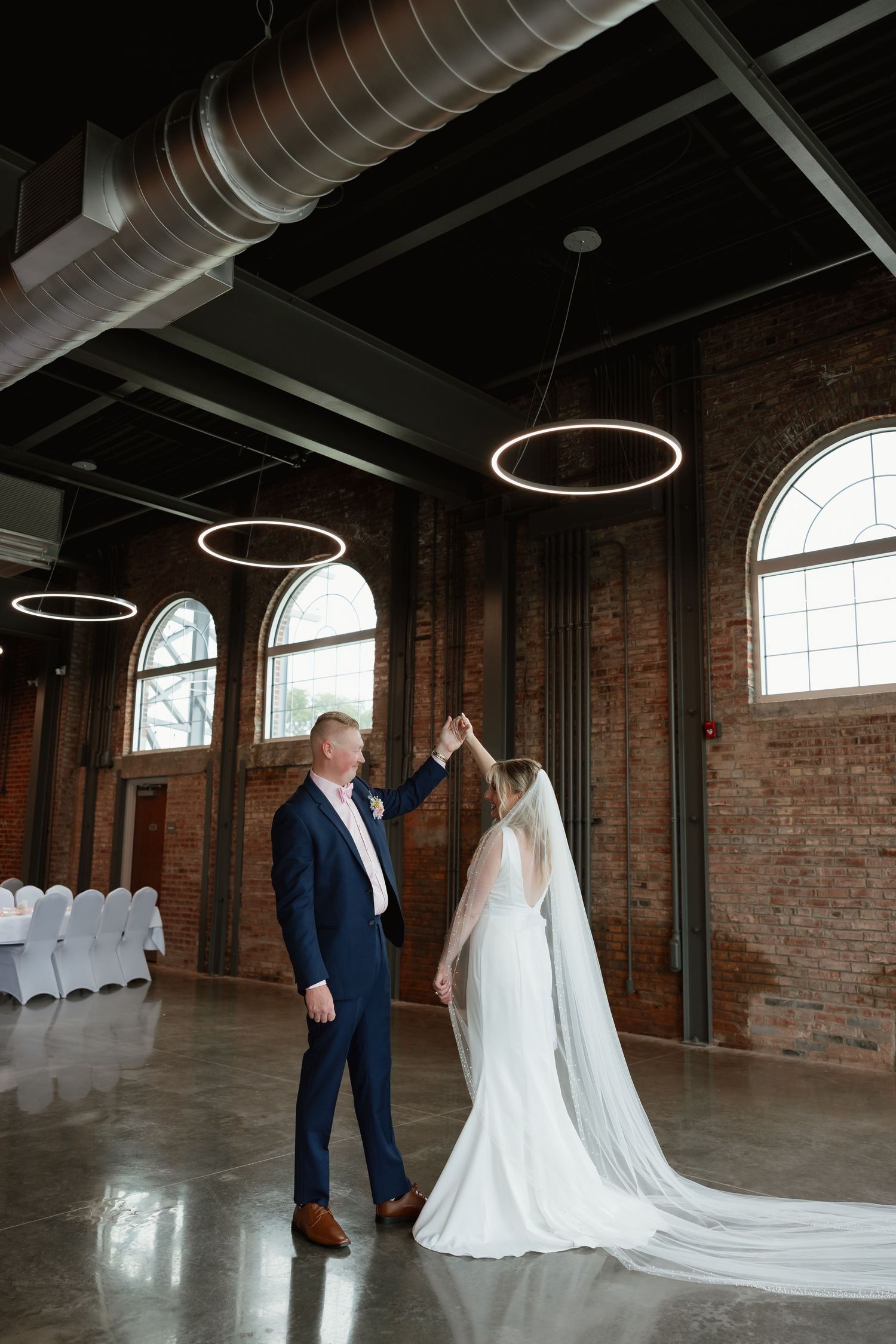 Groom twirls bride in elegant white gown with long veil in stunning industrial brick reception hall.
