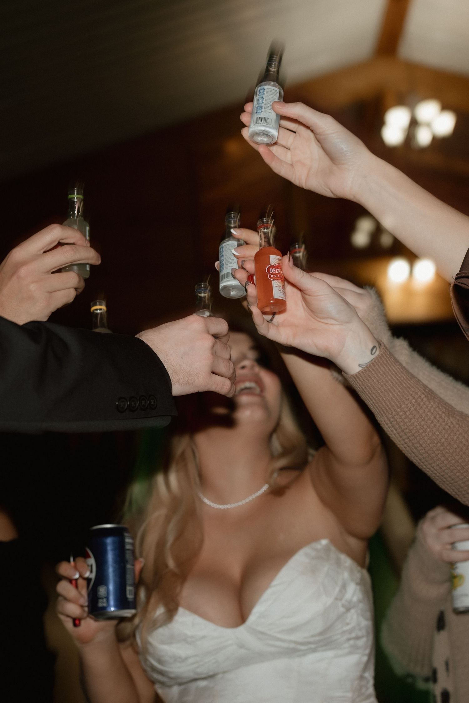Wedding guests toasting with mini liquor bottles while blonde bride laughs at reception.
