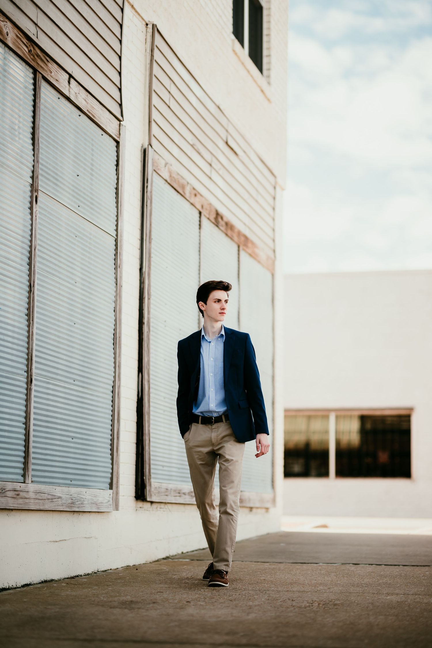 Young man in navy blazer and khakis walks along urban building exterior on sunny day.