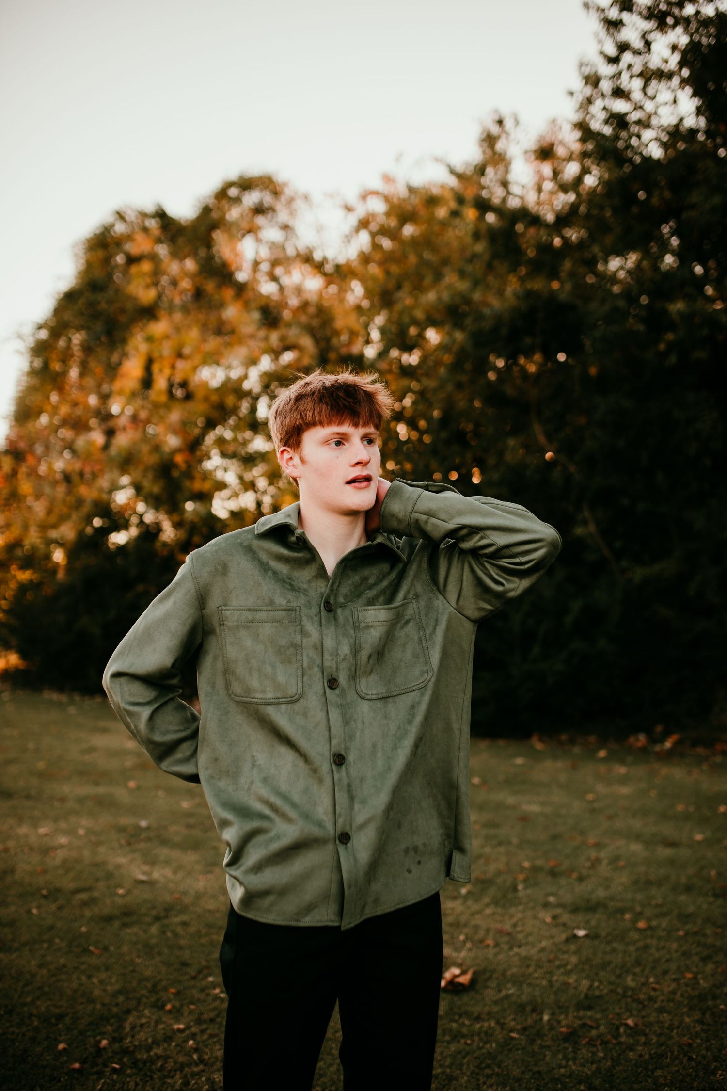 Young man in olive green shirt standing in autumn park at golden hour.