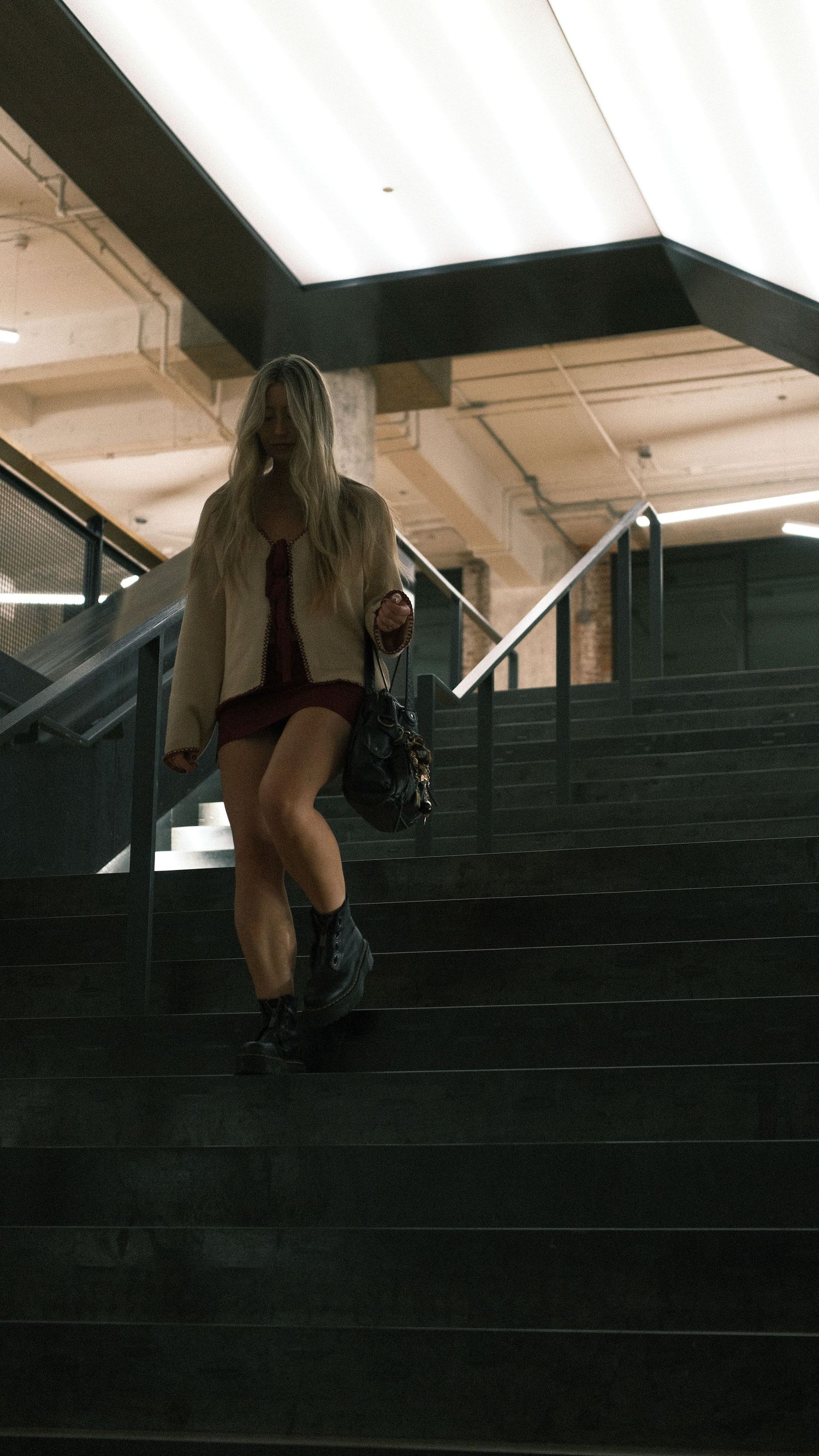 Blonde woman descending dark stairs in red dress, blazer, and black boots.