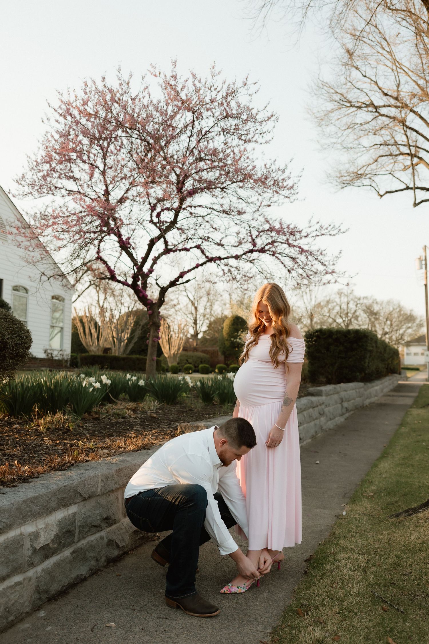 Man kneeling to fix pregnant woman's shoe outdoors in spring with pink blossoming tree backdrop.