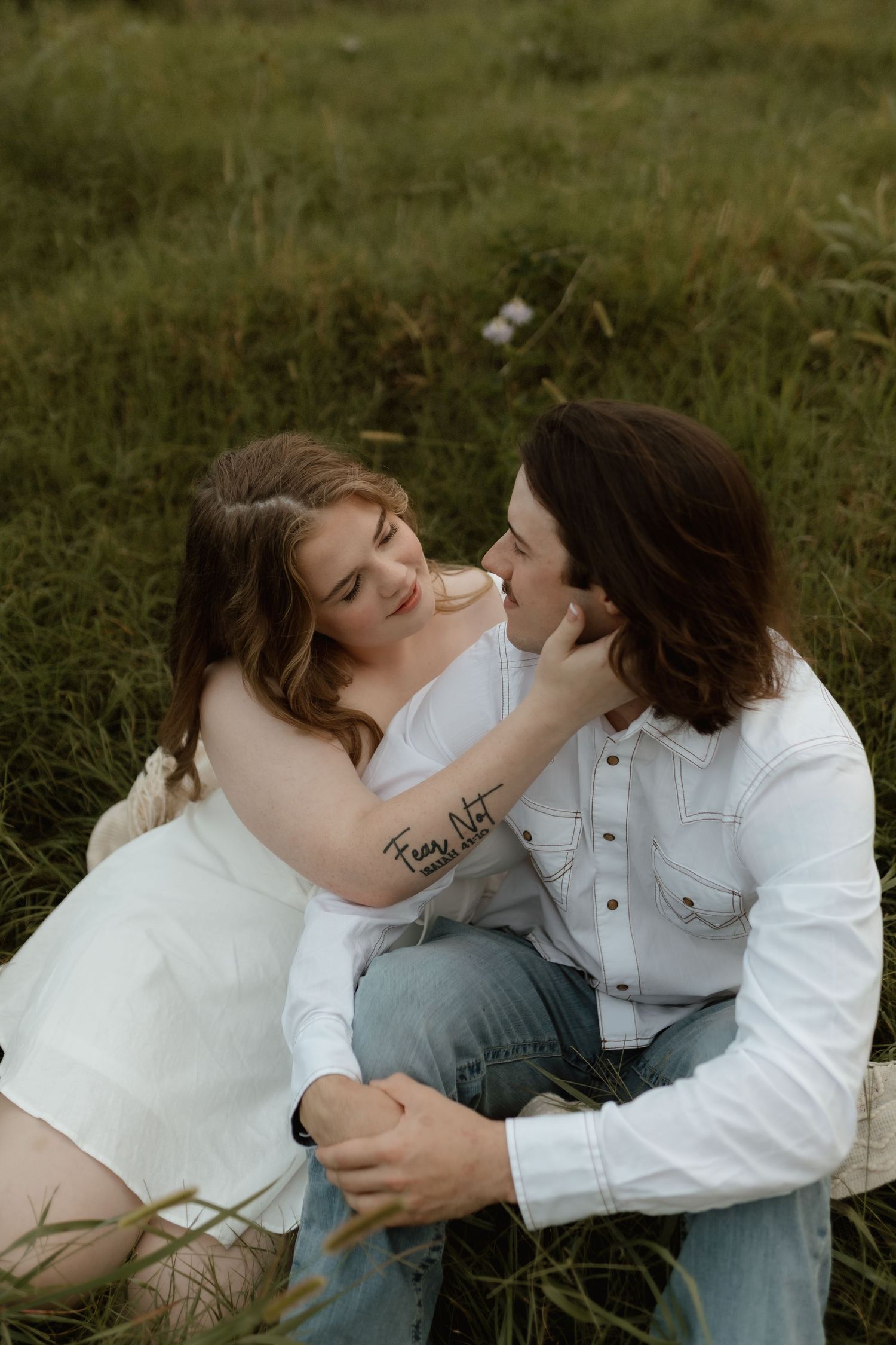 Romantic couple sitting in green meadow grass, woman in white dress touching mans face tenderly at golden hour.