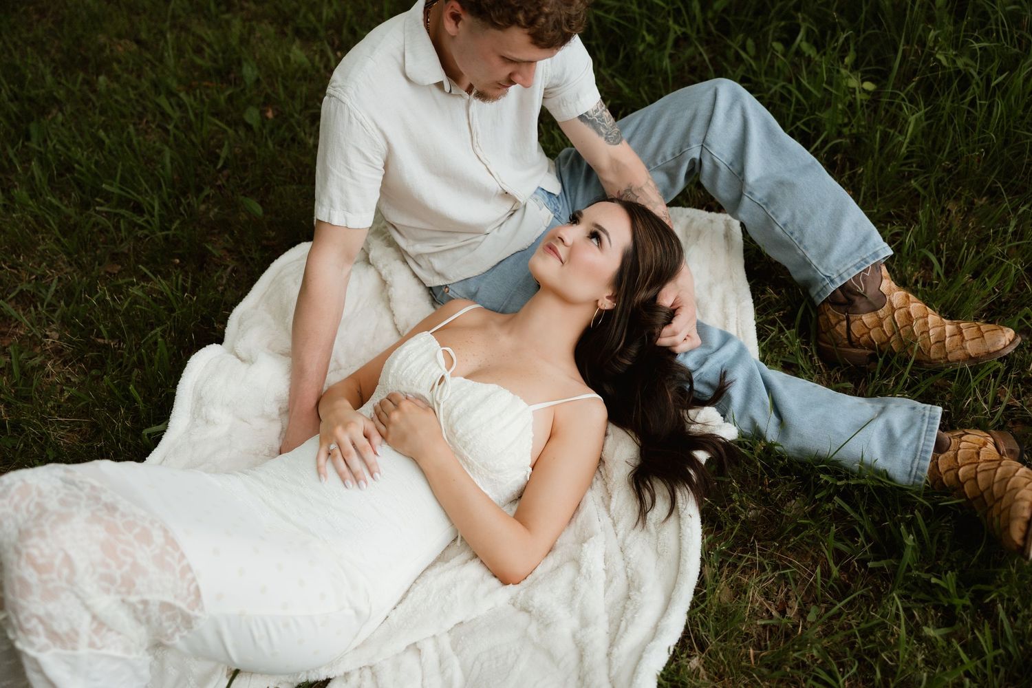 A couple relaxes on the grass as the woman lies back in a white dress outdoors.