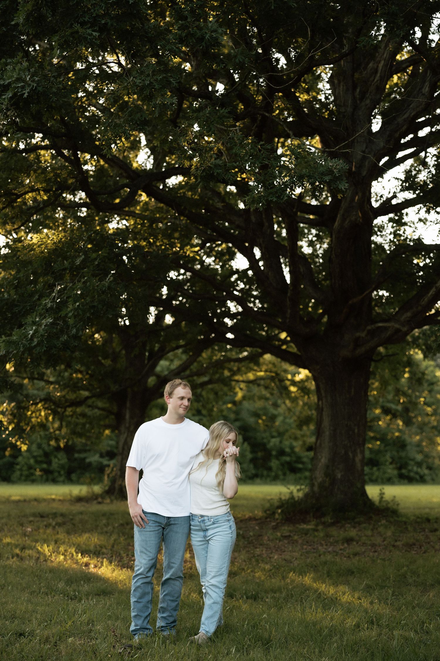 A couple standing together on a grassy lawn beneath two large oak trees in warm golden evening light.