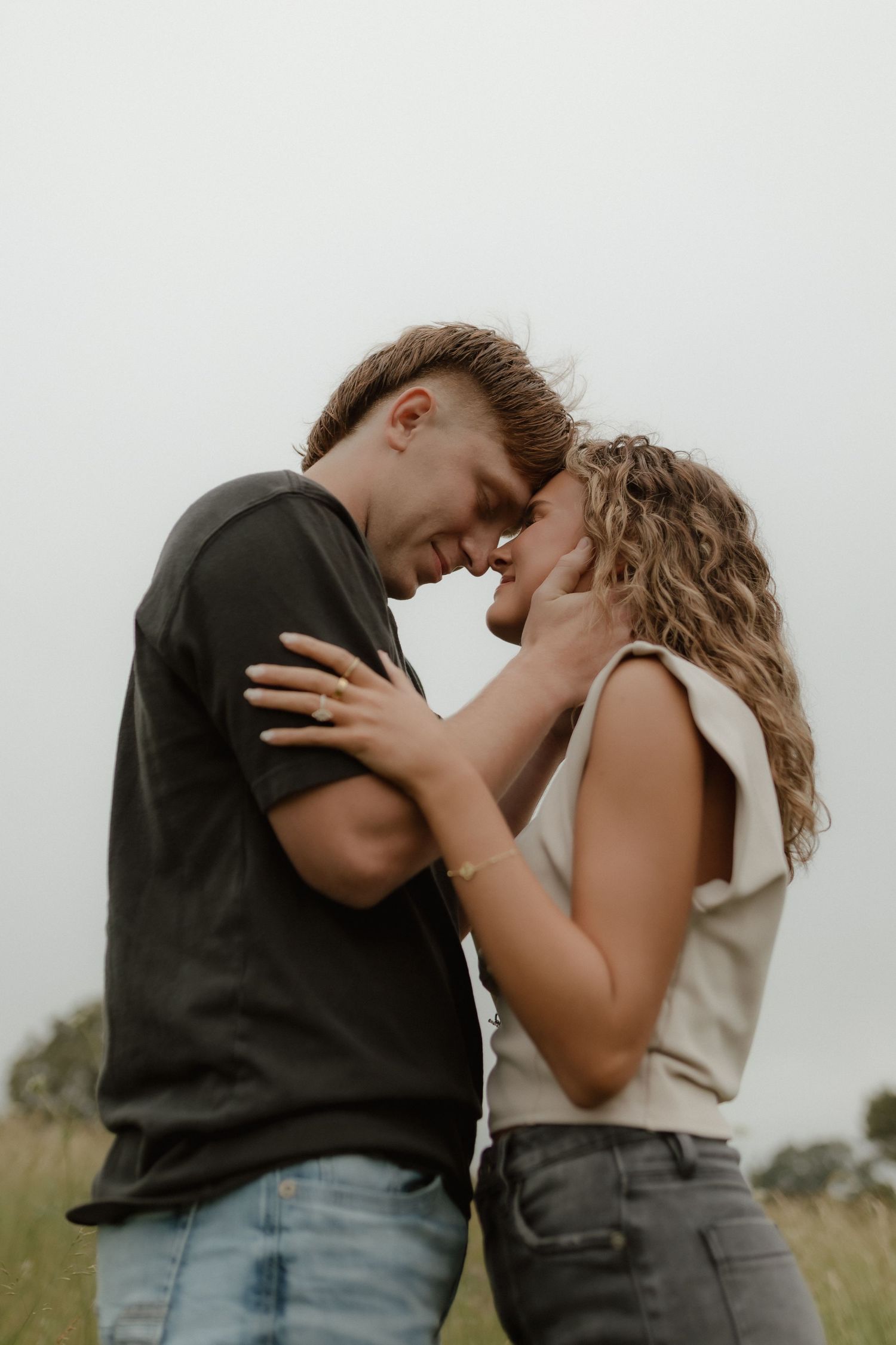 Young couple touching foreheads and smiling in an open grassy field, sharing a tender romantic moment.