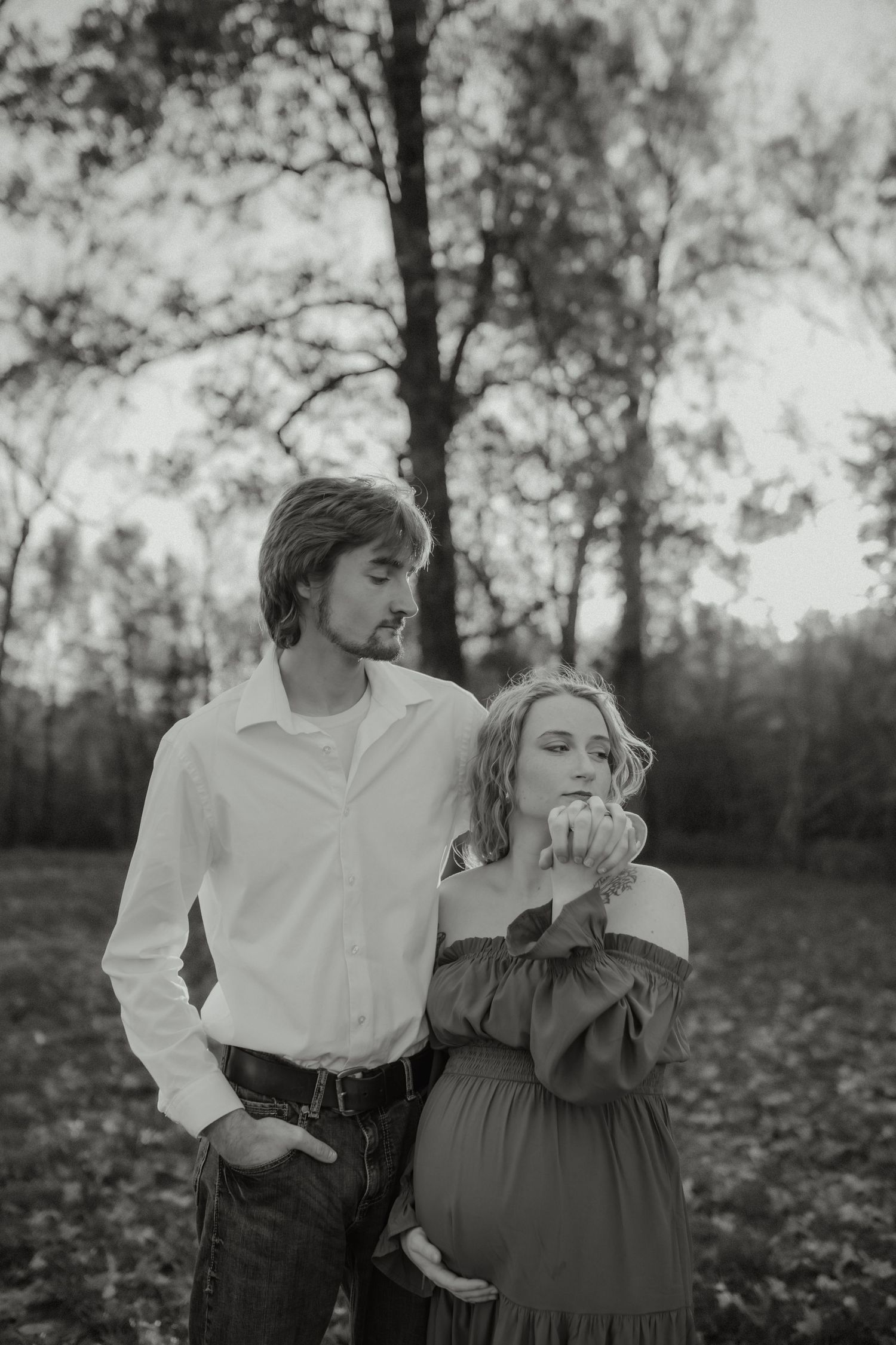 Black and white photo of couple in fall forest, pregnant woman in off-shoulder green dress.