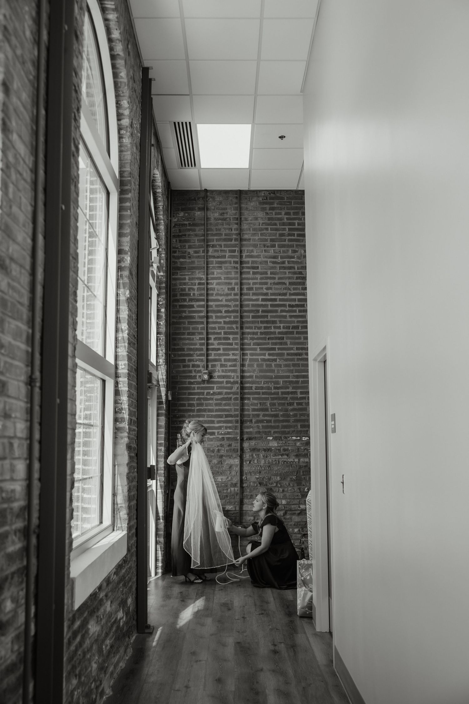 Bride gets veil adjusted by assistant in a narrow brick-walled corridor in black and white photo.