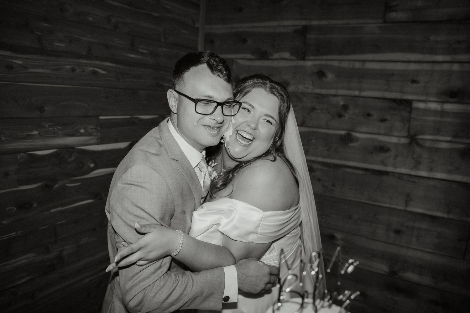 Happy bride and groom embracing and smiling together against rustic wooden barn wall.