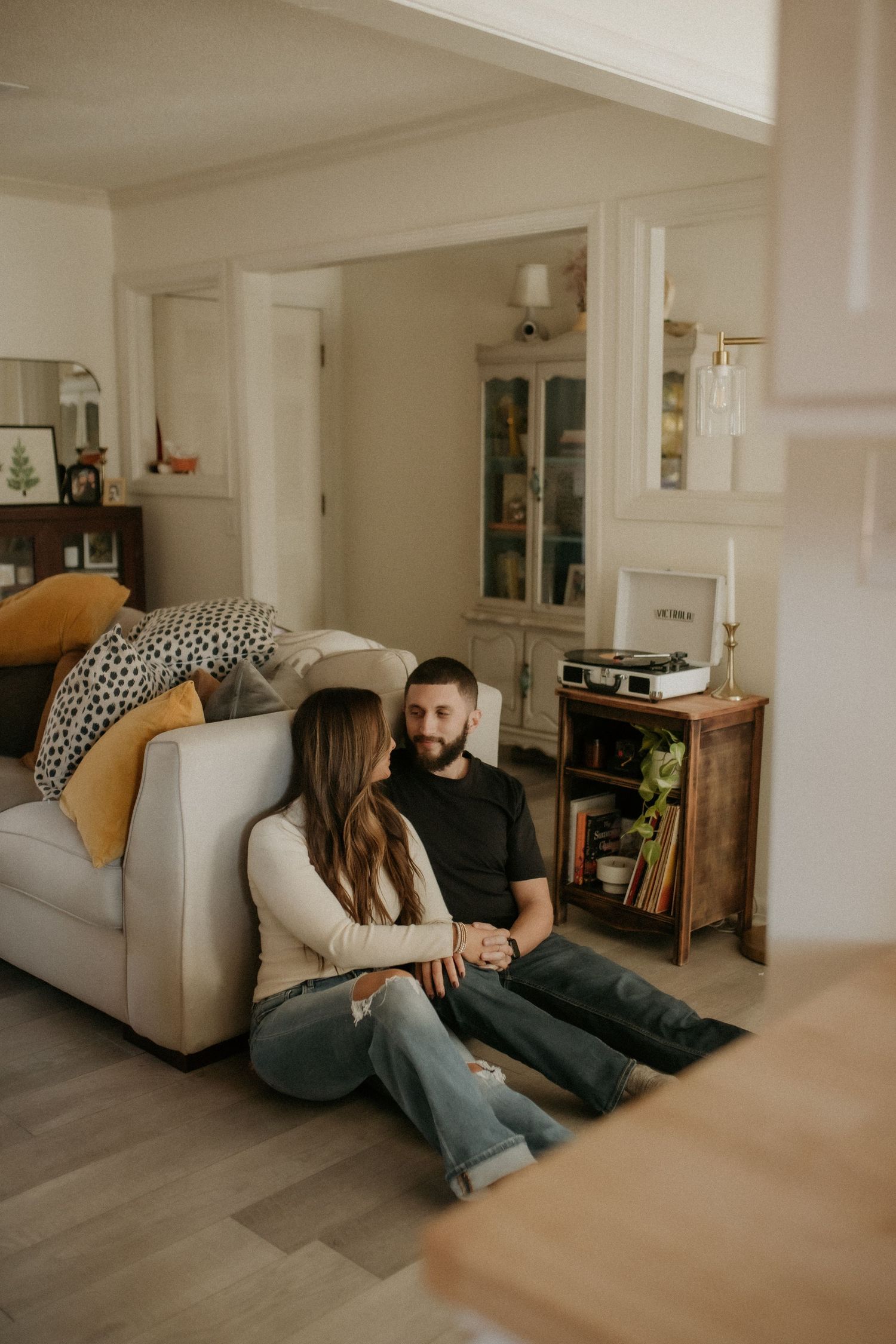 Couple sits together on living room floor, holding hands and sharing a quiet intimate conversation at home.