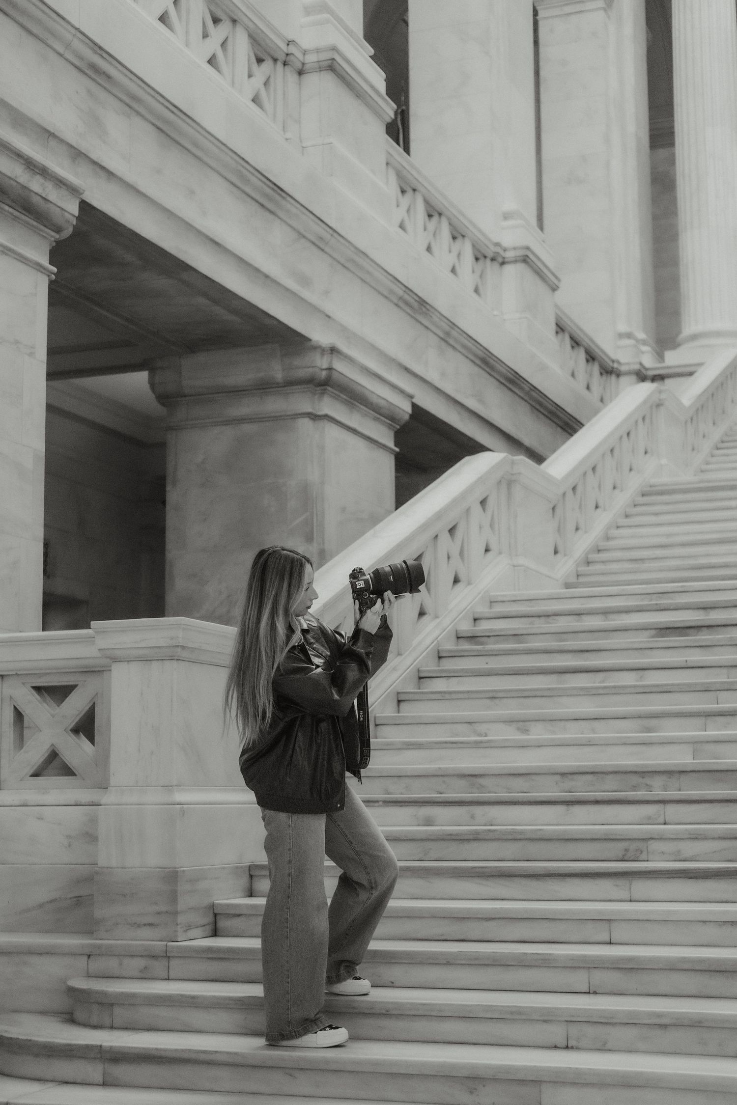 Woman with long hair photographing marble staircase of classical building, black and white.