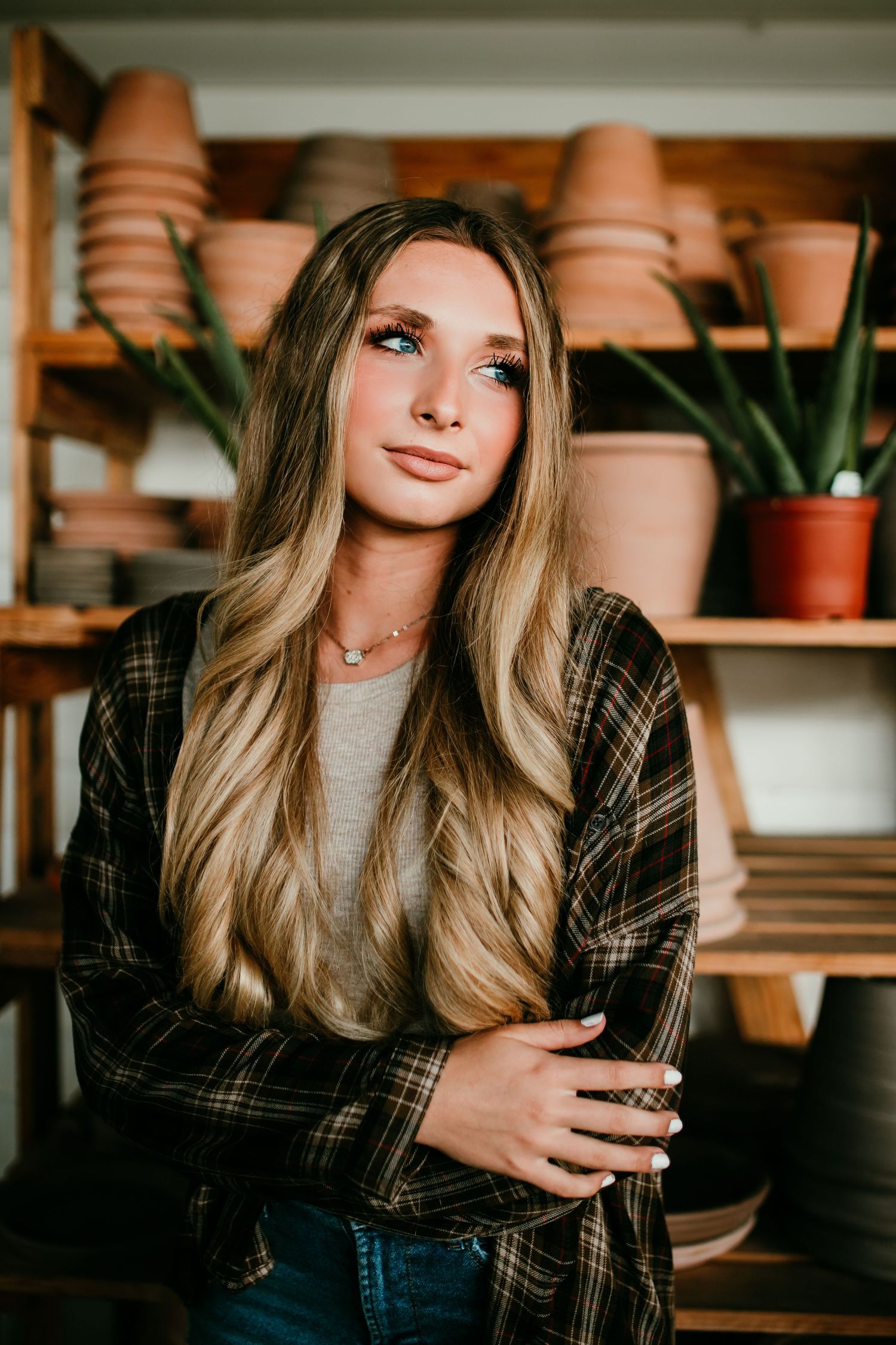 Young woman with long blonde hair posing in plaid jacket among terracotta pots and plants.