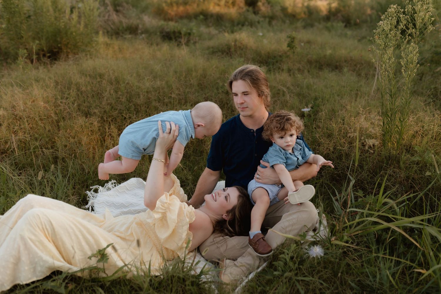 Family relaxing in grassy field at golden hour, mother lying down lifting baby while father holds toddler.