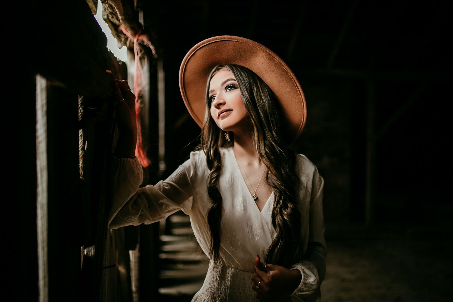Brunette woman in wide-brim hat and white blazer poses dramatically in moody dark setting with warm light.