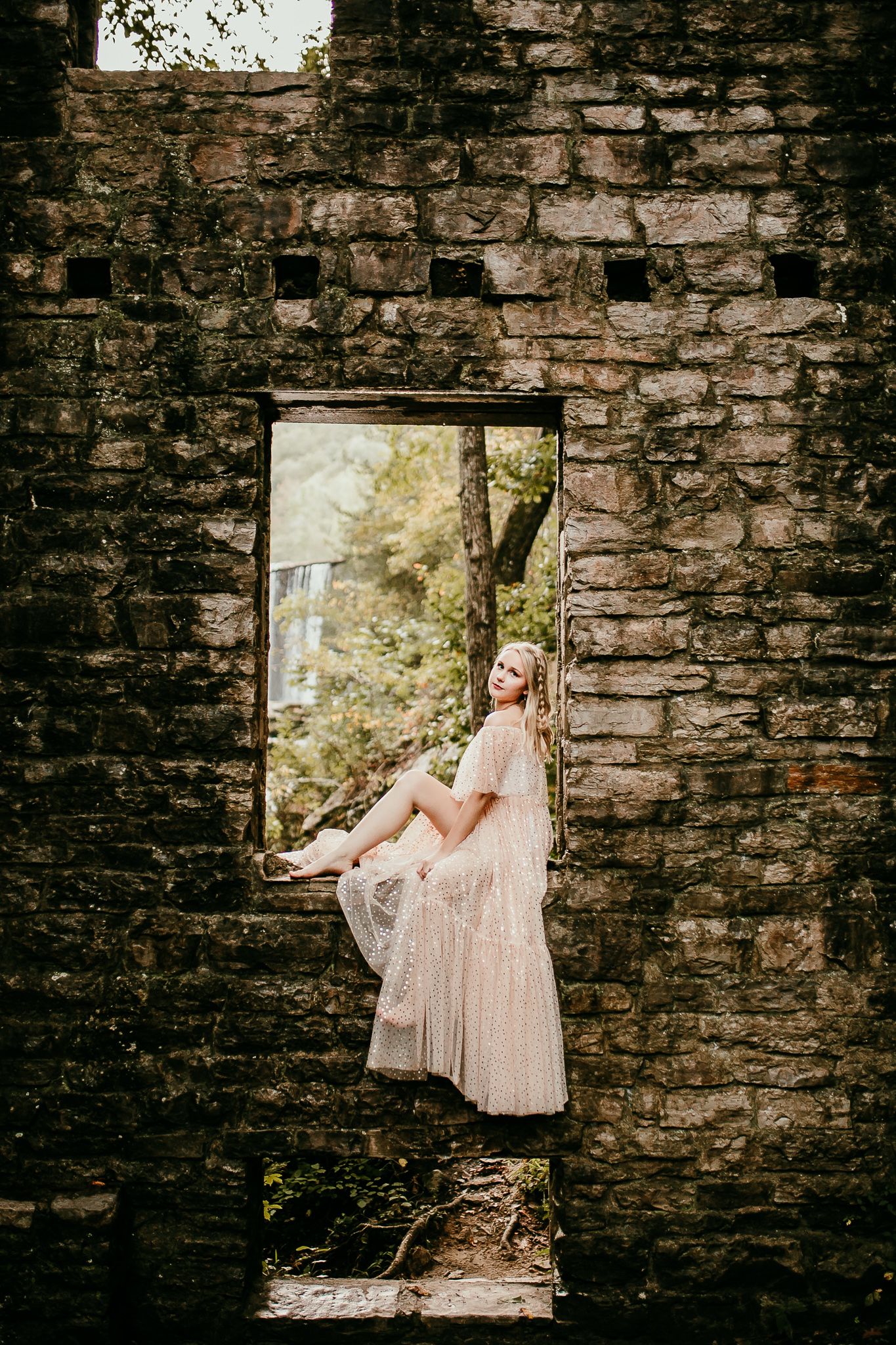 Woman in flowing sparkly blush gown sits in window opening of rustic stone ruins surrounded by greenery.