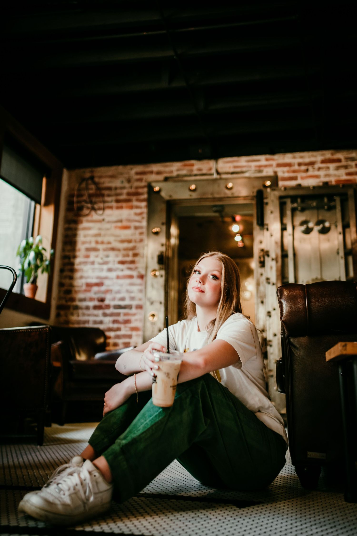 Blonde teenage girl sits on floor against leather chair in rustic brick-walled coffee shop.