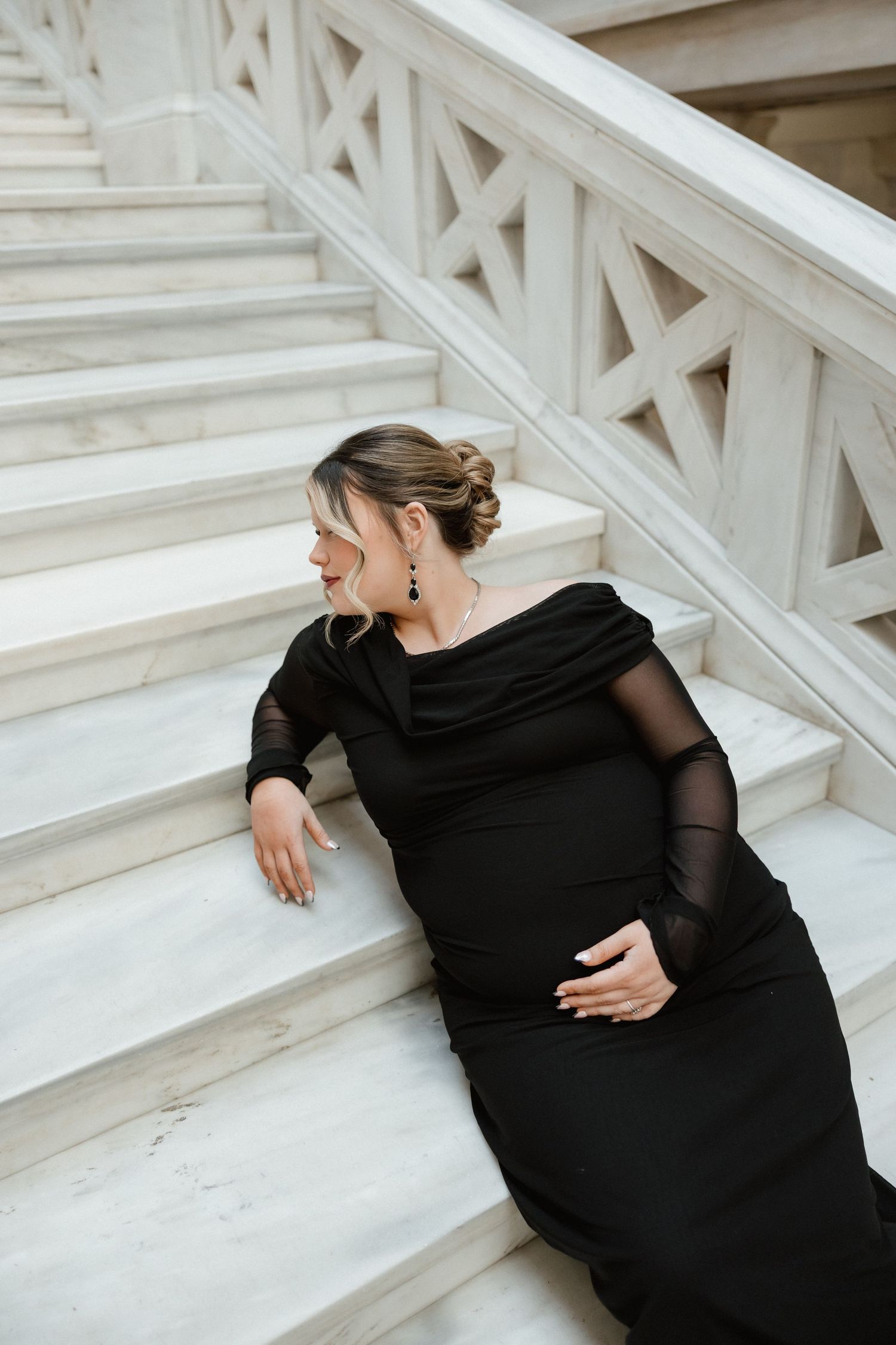 Pregnant woman in black off-shoulder gown sitting gracefully on white marble staircase steps.