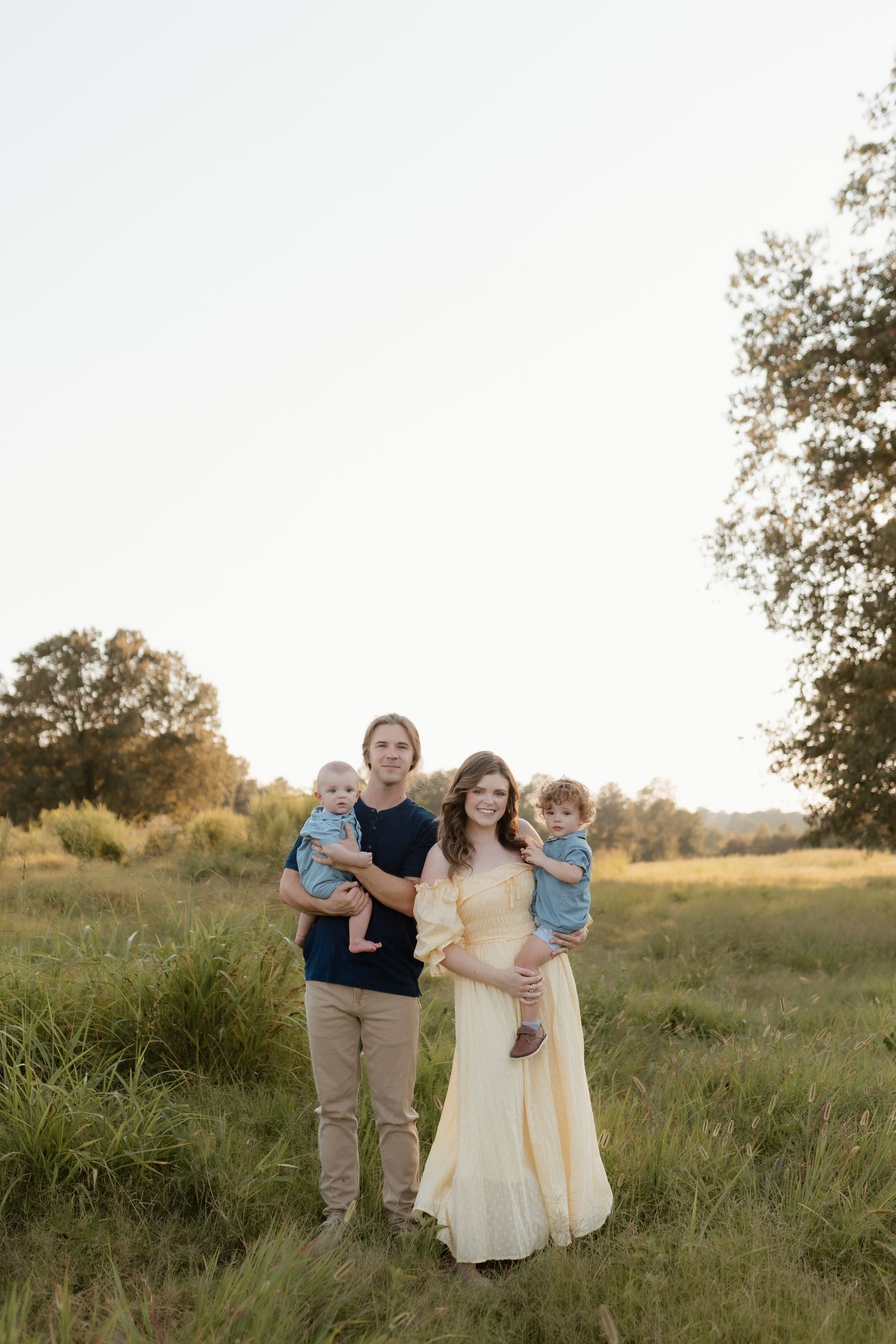 Family of four with two young children poses together in golden sunlit grassy meadow.