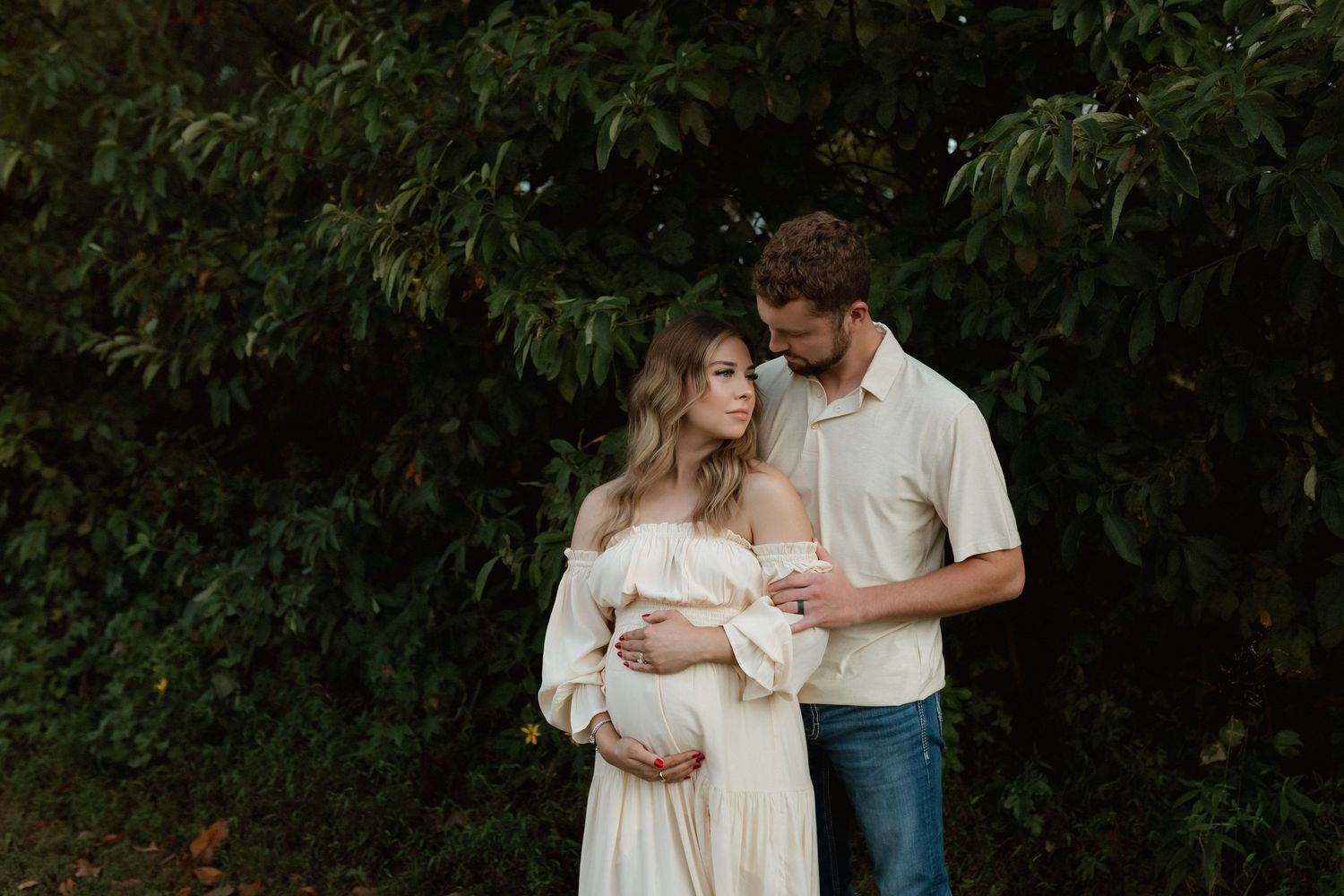 A couple tenderly embracing outdoors in front of lush green trees.