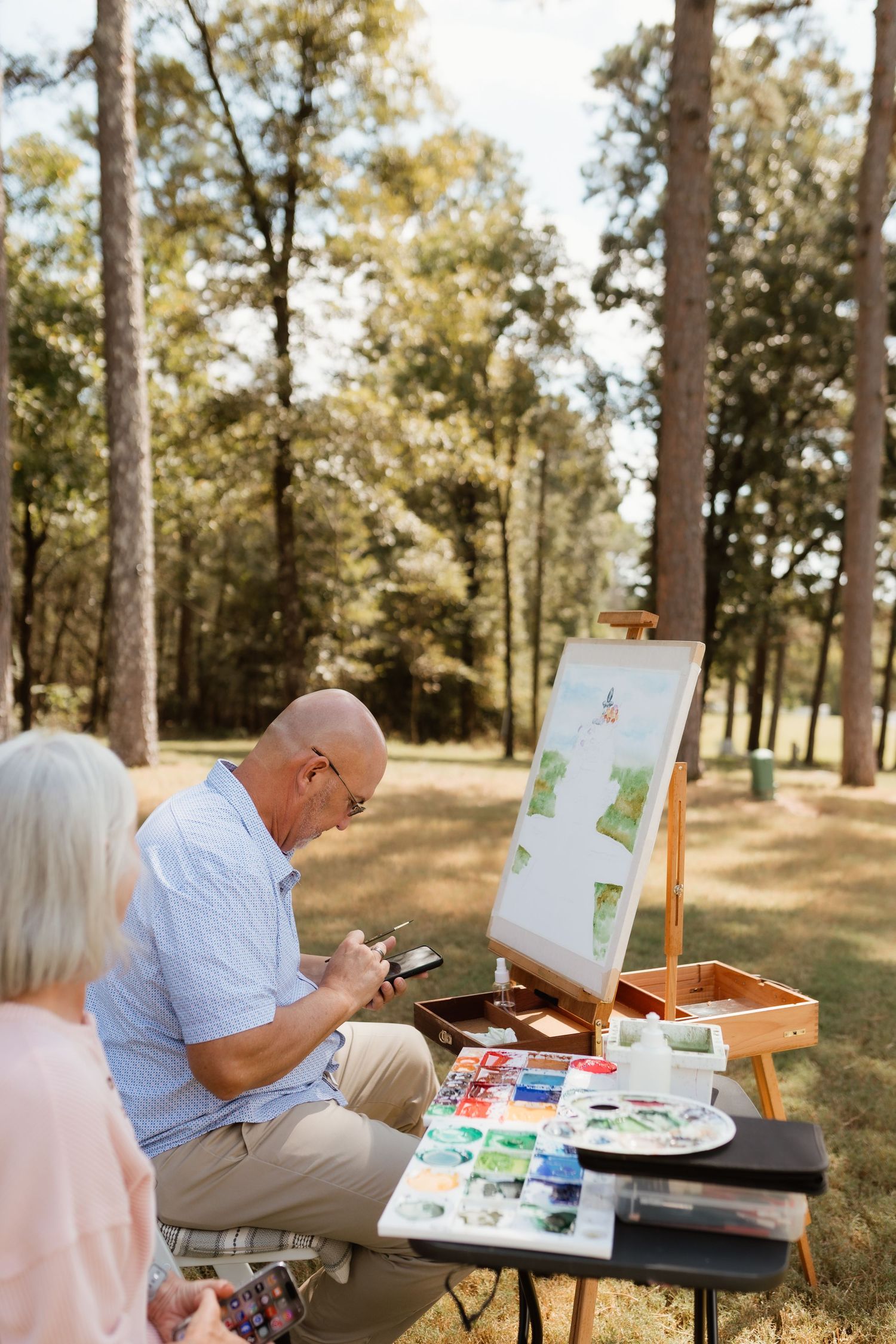 Elderly couple painting watercolors on an easel outdoors in a sunny forest park setting.