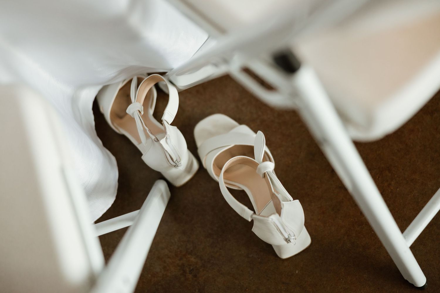White bridal sandals with ankle straps resting on a brown floor between white chairs at a wedding venue.
