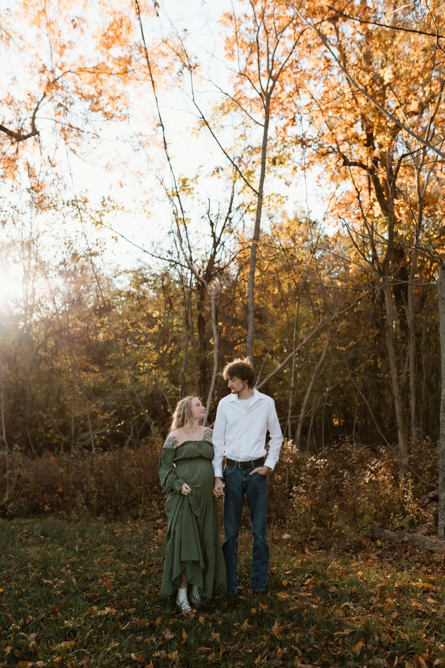 Expecting couple holding hands and gazing at each other among vibrant autumn trees in golden light.