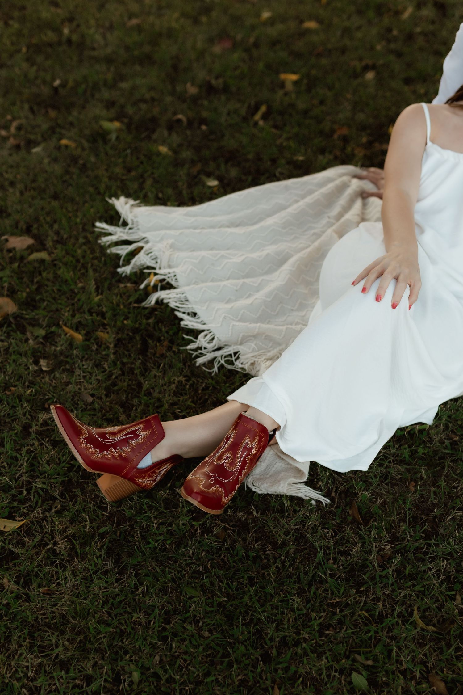 Close-up of a woman in a white dress and red cowboy boots sitting on green grass with a fringe blanket.