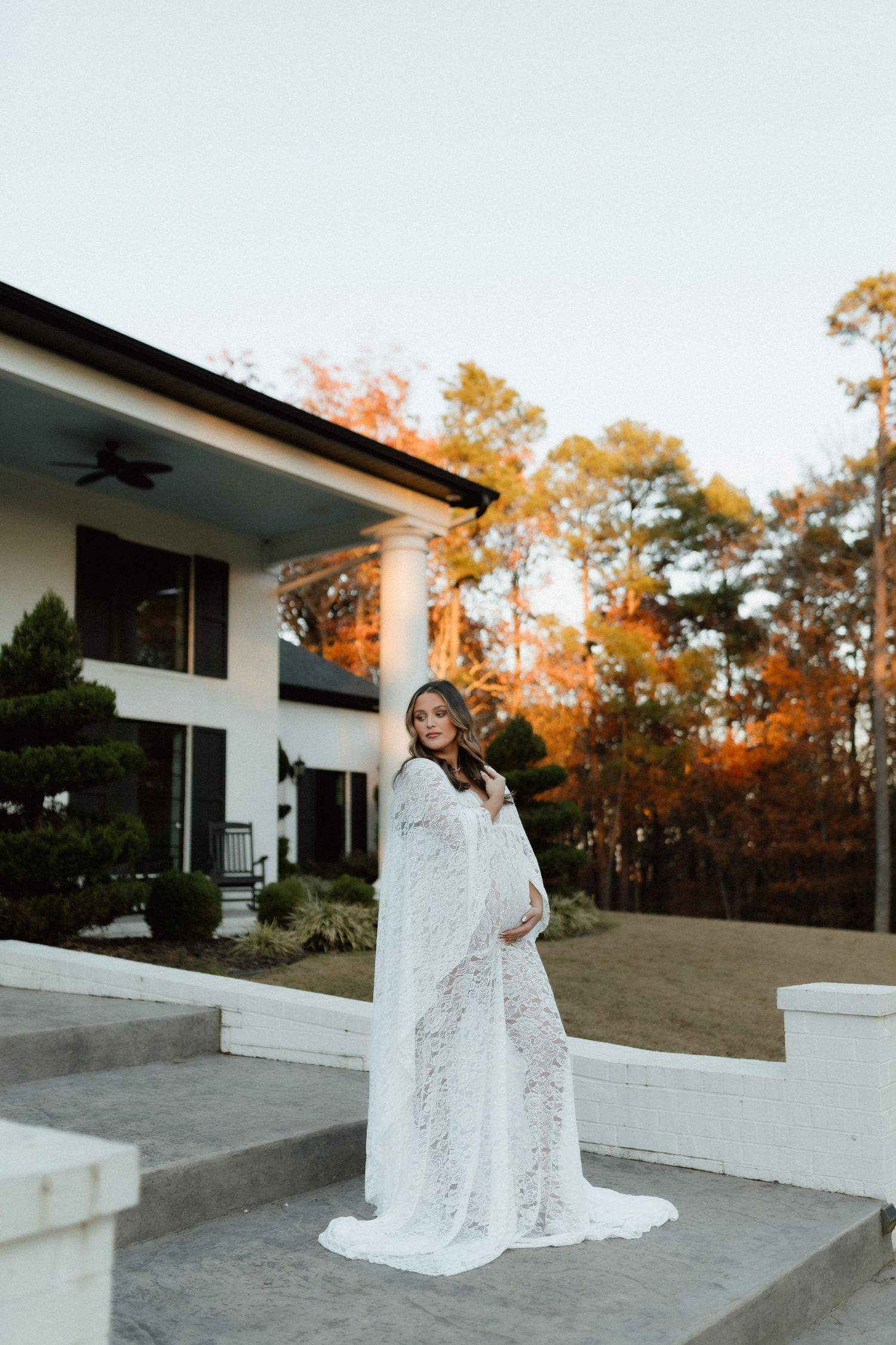 Pregnant woman in white lace maternity gown standing on steps outside white house with autumn foliage.