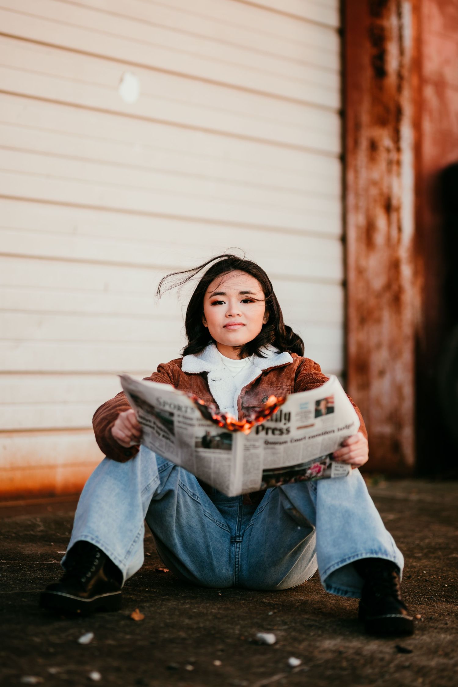 Young Asian woman sitting on ground holding a burning newspaper wearing brown jacket and jeans outdoors.