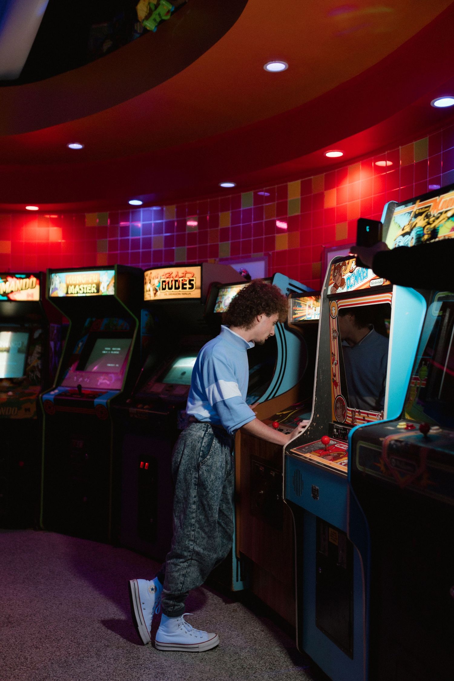 Young man in retro outfit playing classic arcade game in vibrant neon-lit game room.