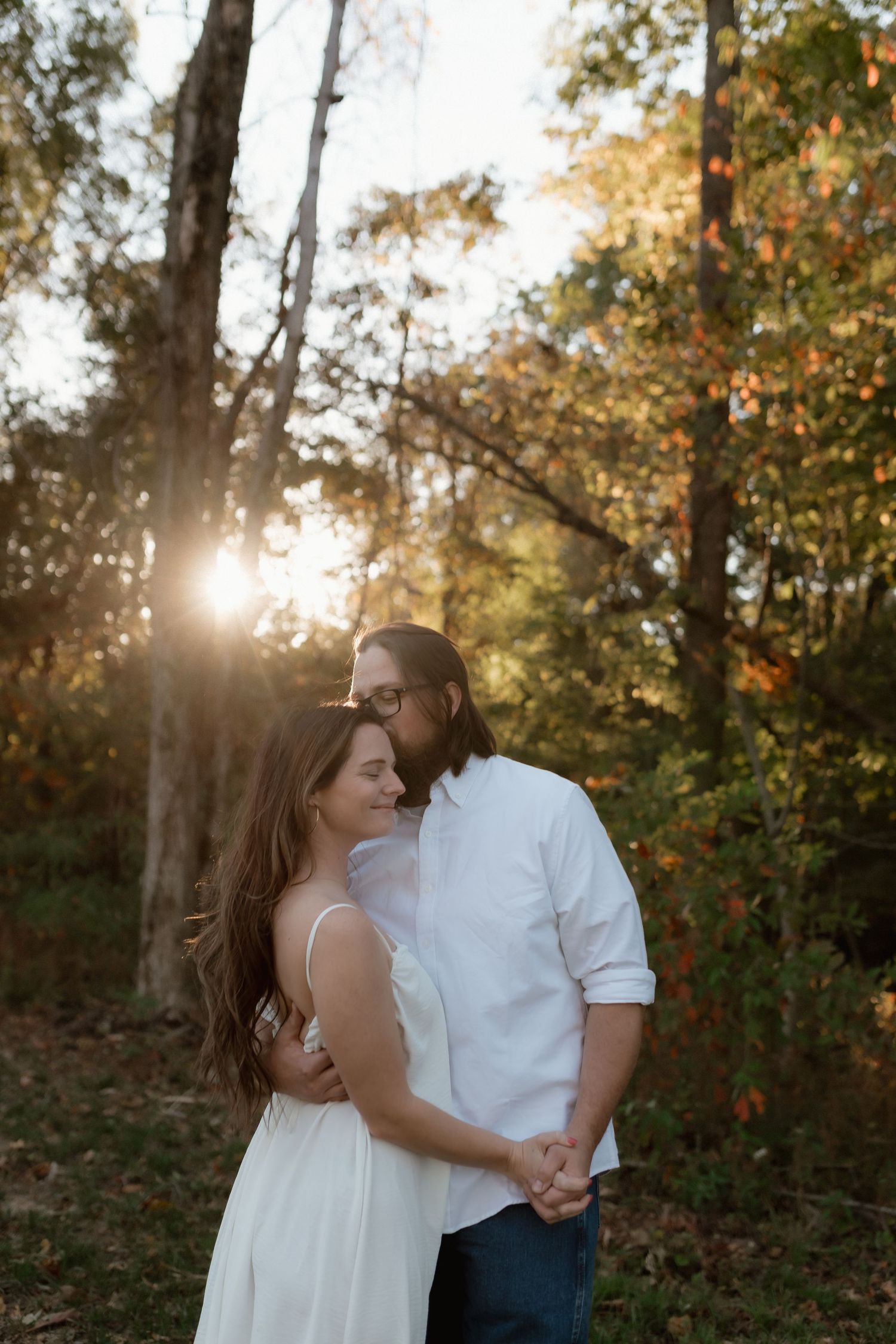 A couple shares a romantic moment in a sunlit forest, man kissing womans temple as golden light streams through trees.