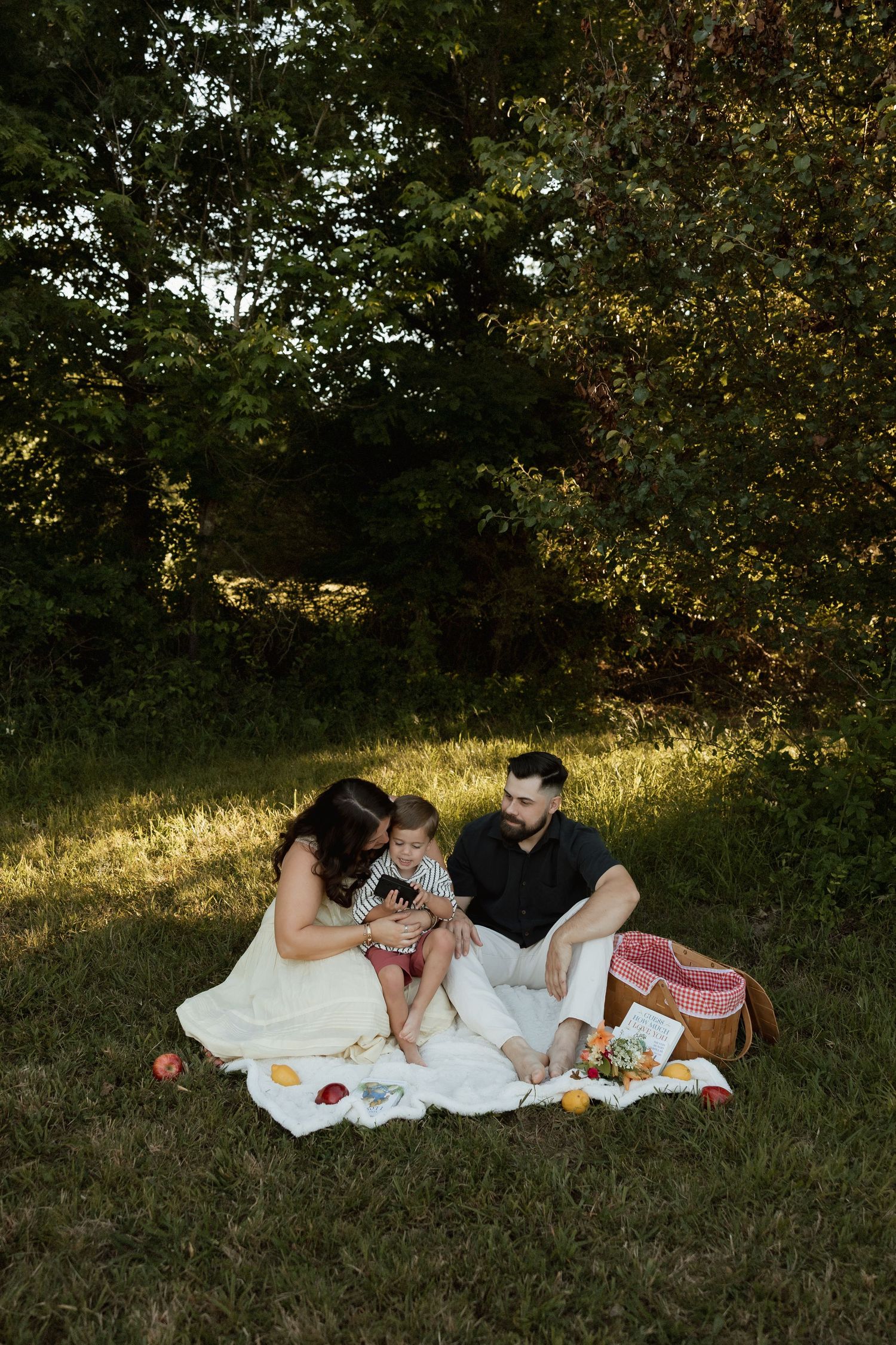 Family sitting on white blanket reading book together during outdoor picnic in dappled sunlight.