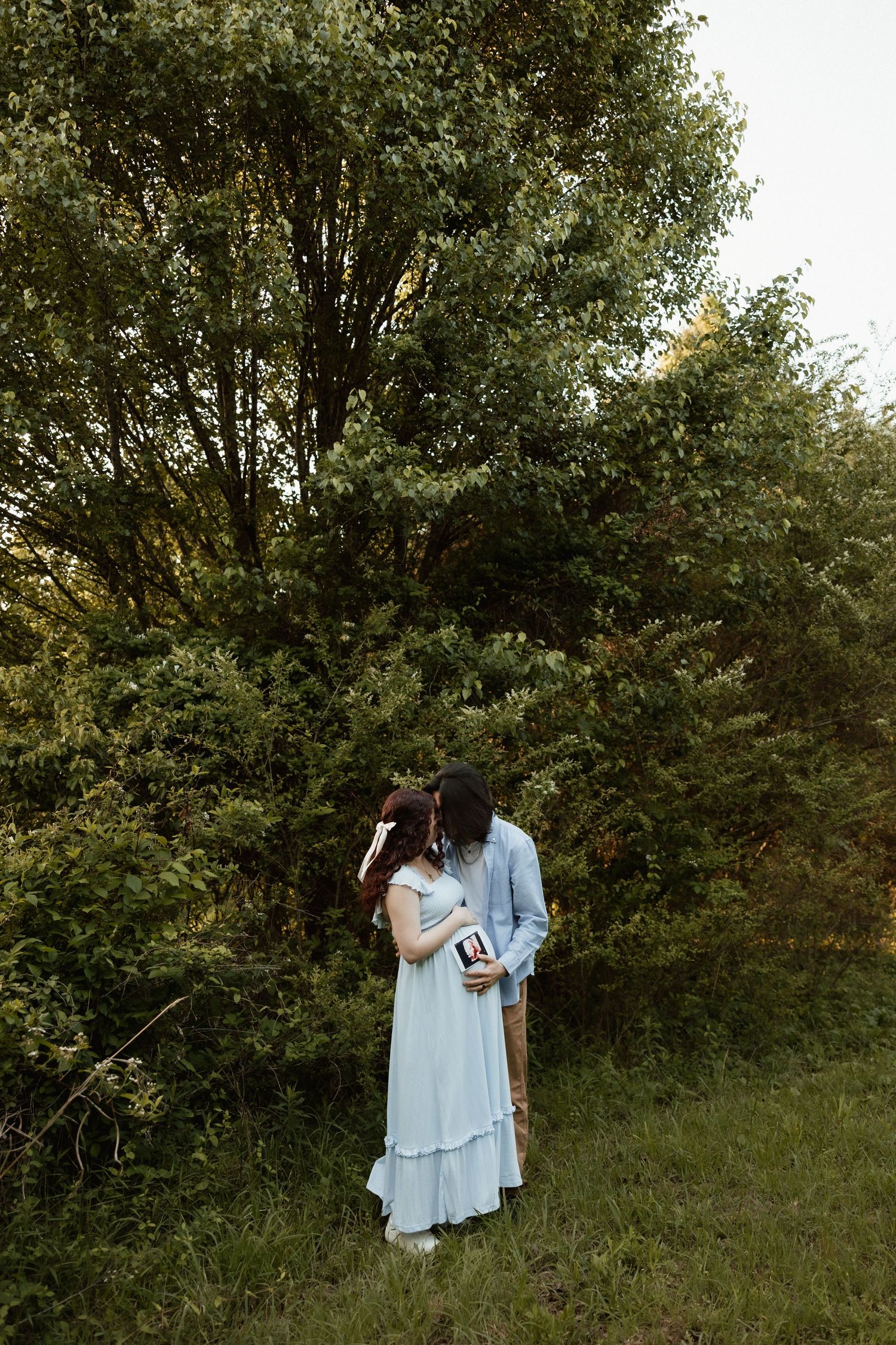 Expecting couple embracing and kissing while holding ultrasound photos in a green forest setting.