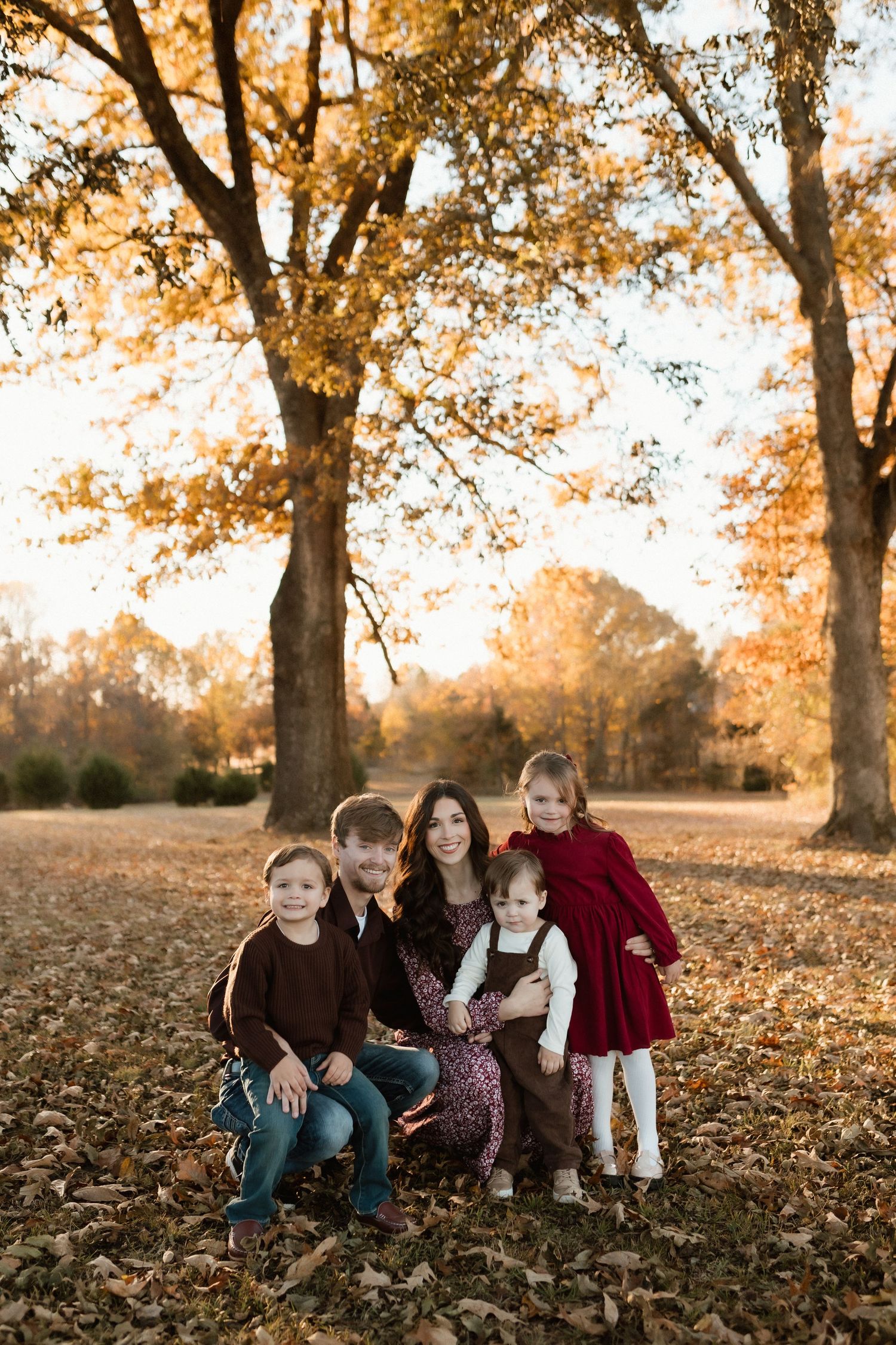 Family of five poses together on fallen autumn leaves under golden trees during a fall outdoor photo session.