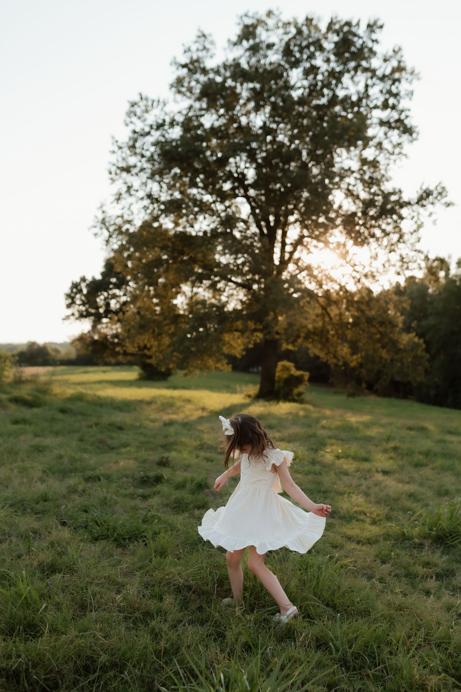 Young girl in white dress twirling in a sunlit green field with a large tree glowing at golden hour.