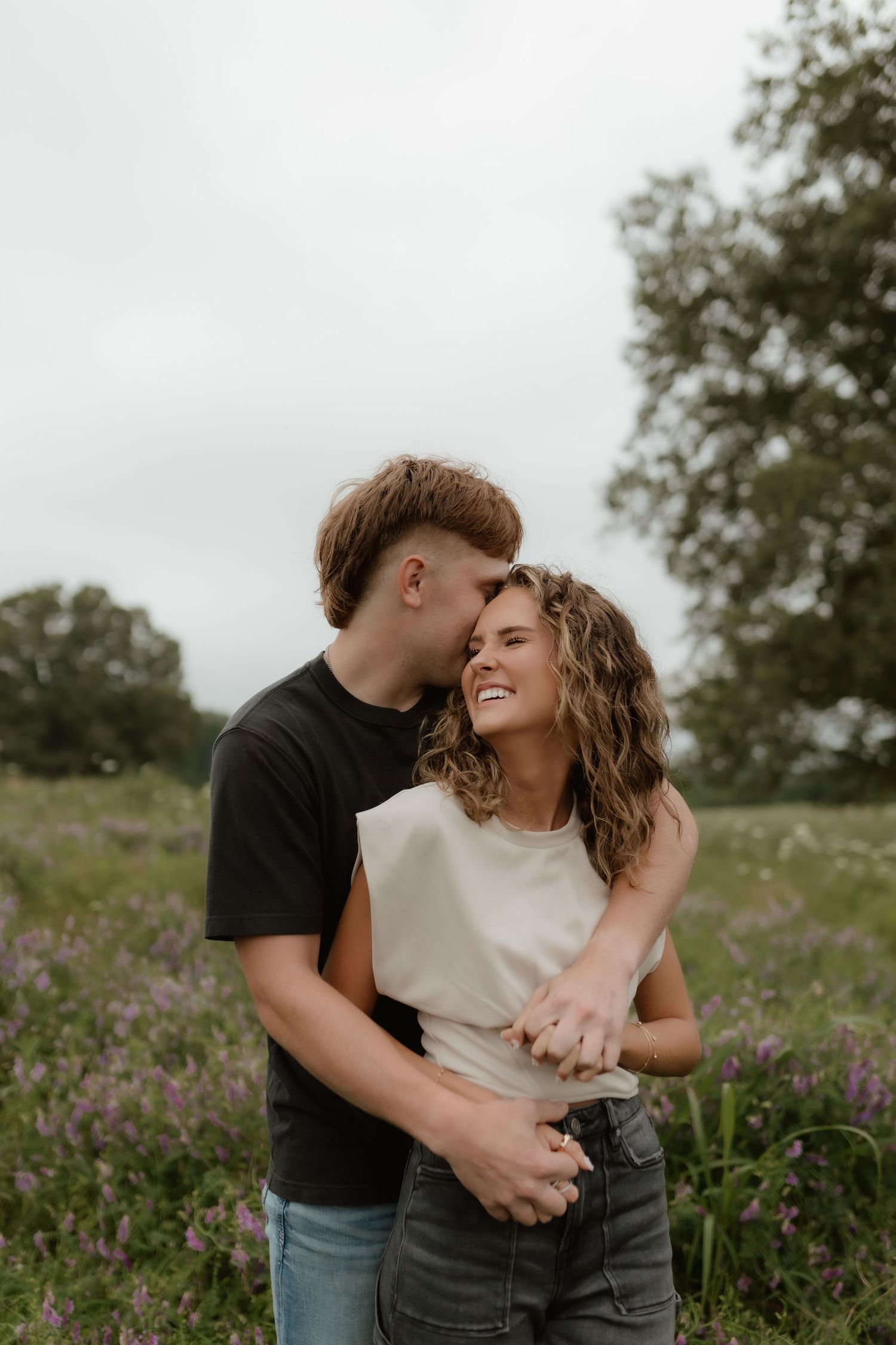 Couple embracing and laughing in a wildflower field, man whispering in womans ear during outdoor engagement session.