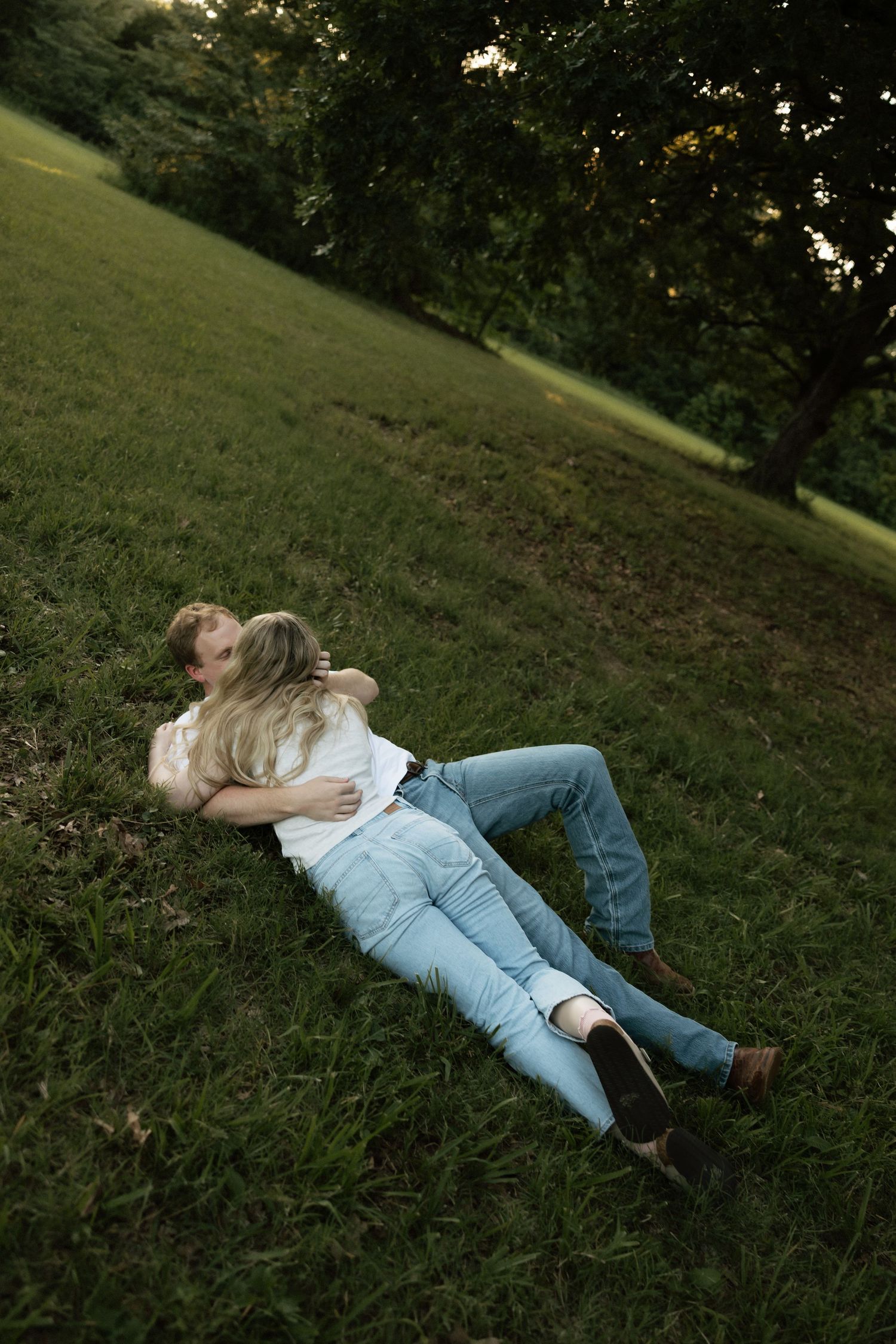 A romantic couple lying together on a green grassy hillside, embracing under shady trees at dusk.